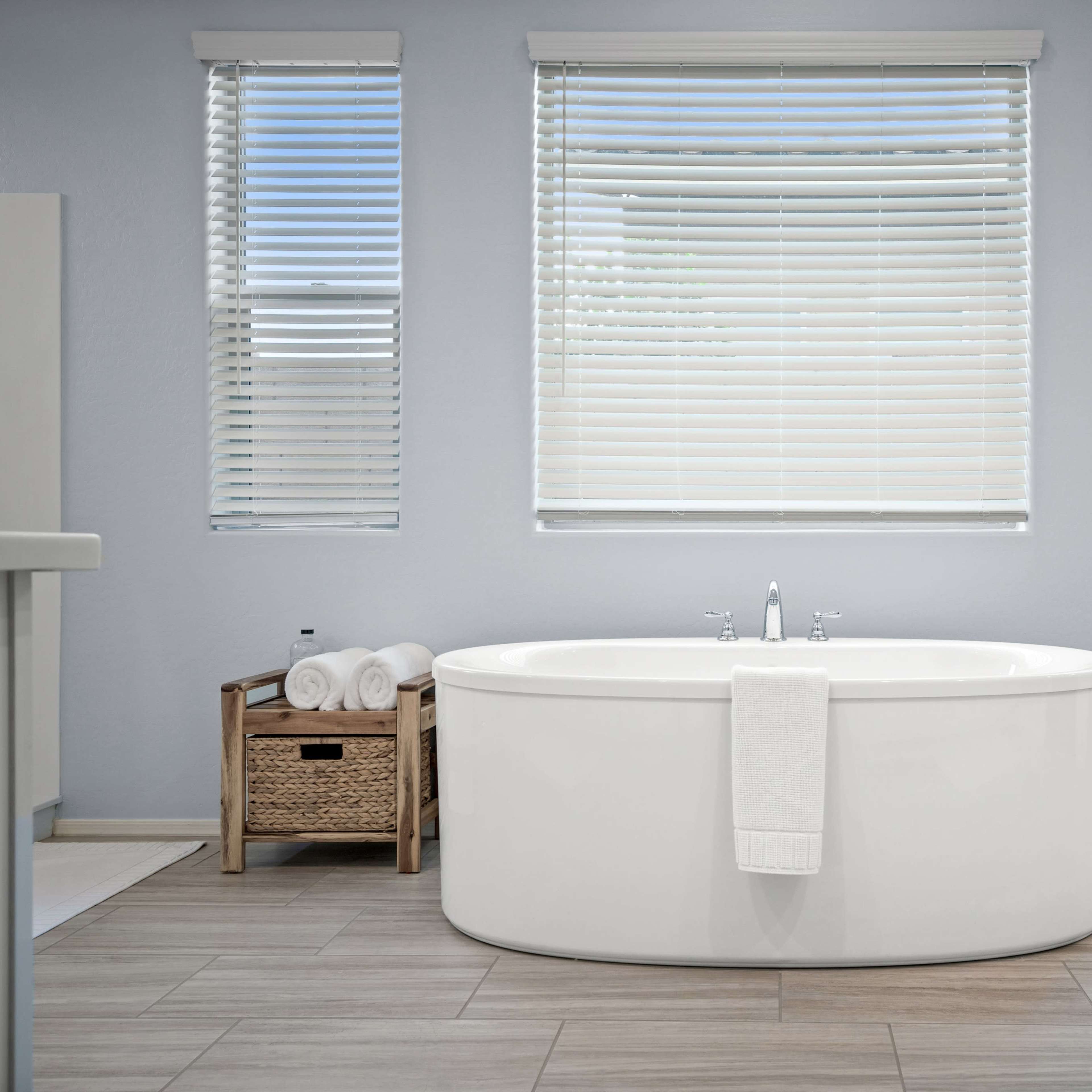 A modern bathroom with a freestanding oval bathtub, two windows with blinds, and a basket containing rolled towels.