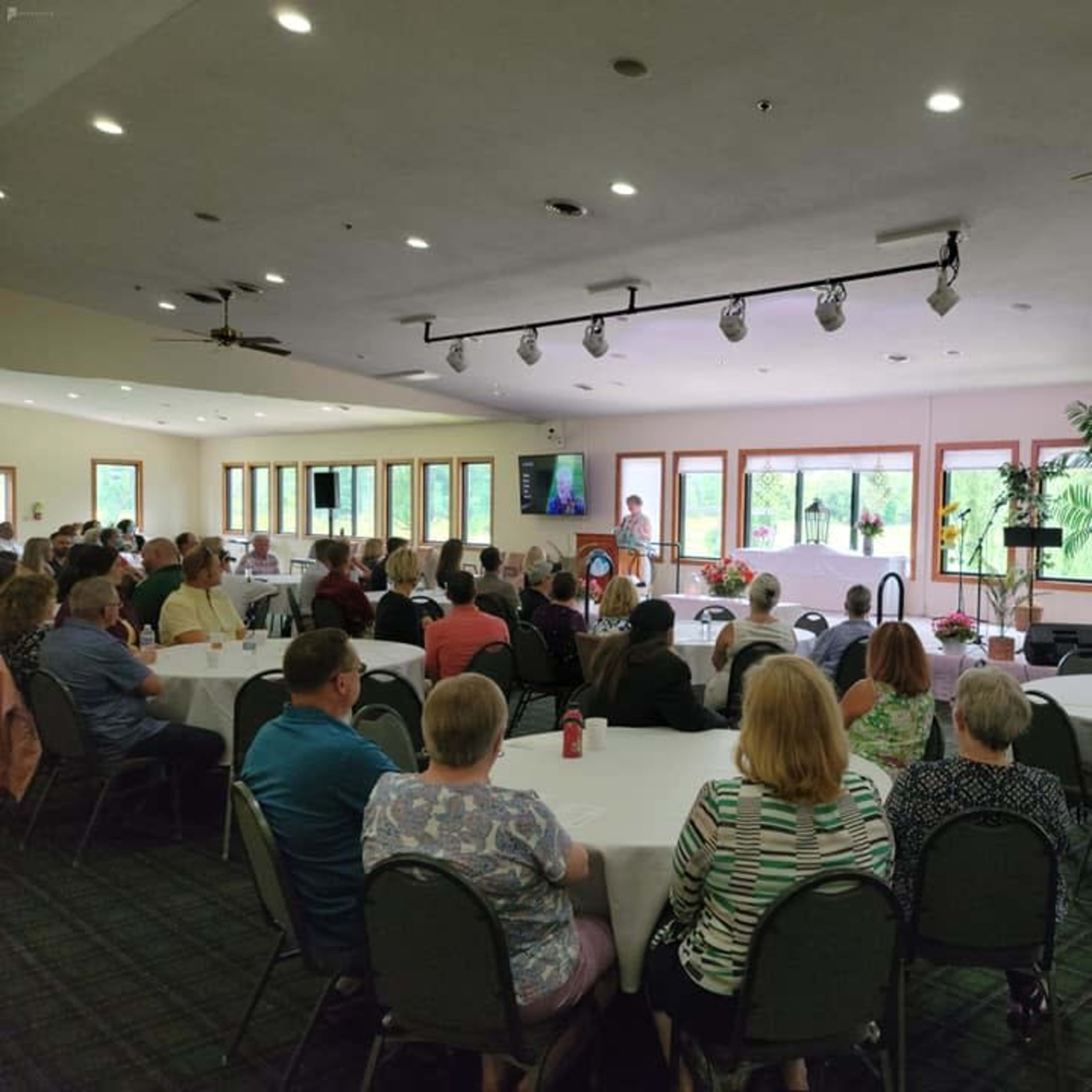 A speaker addresses a large audience seated at round tables in a well-lit conference room with large windows and a stage.