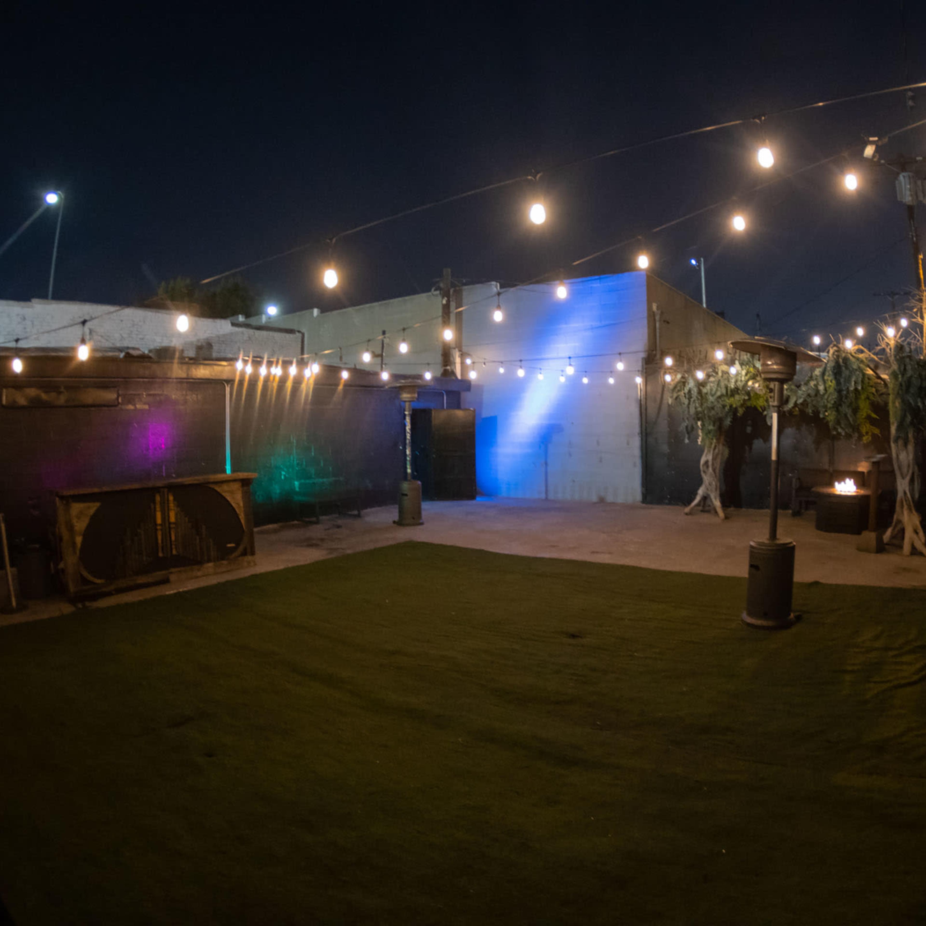 A well-lit outdoor space with artificial grass, string lights overhead, and a fire pit in the corner.