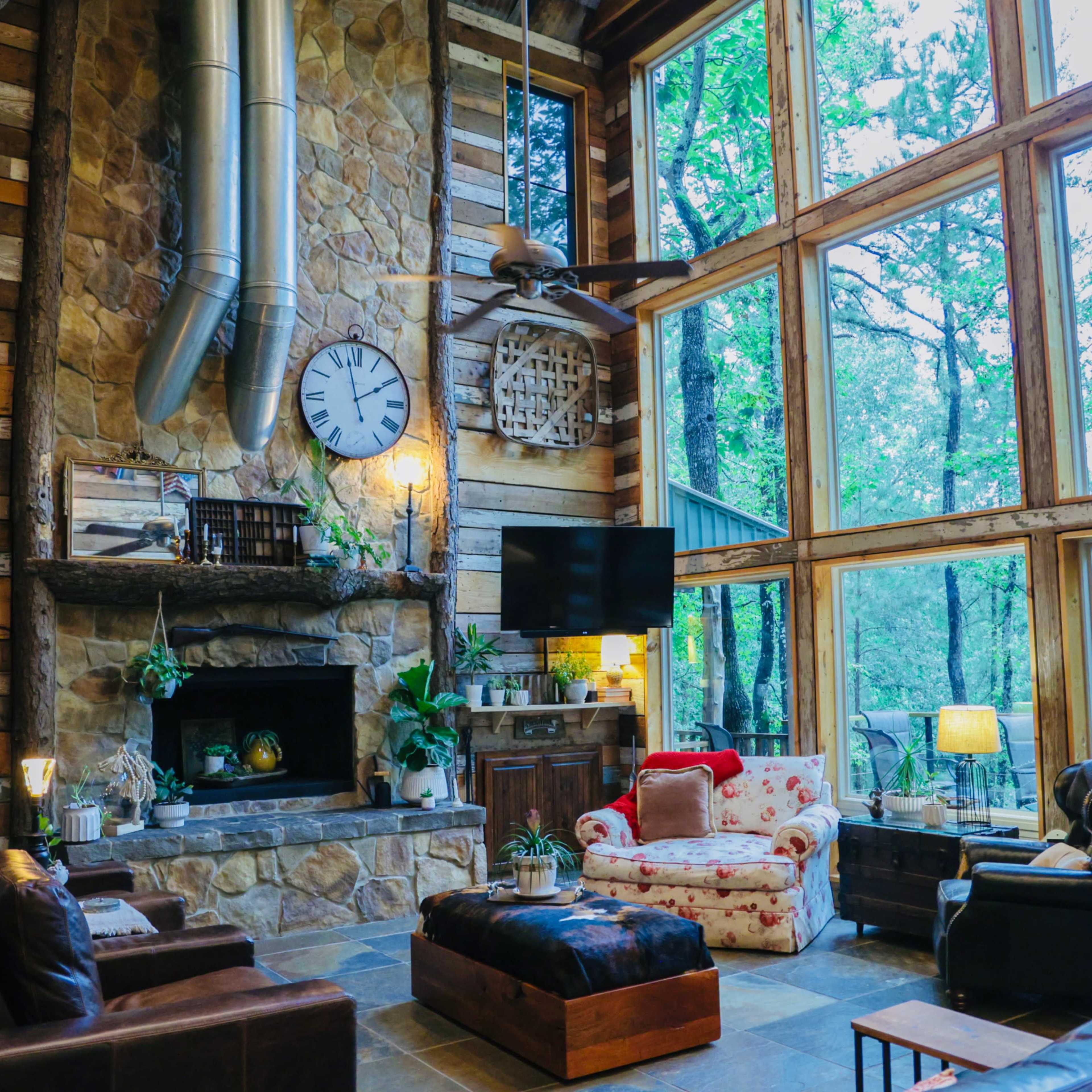 A cozy living room with a stone fireplace, large windows overlooking trees, and rustic wooden walls, featuring a mix of furniture including a floral chair and a dark leather sofa.