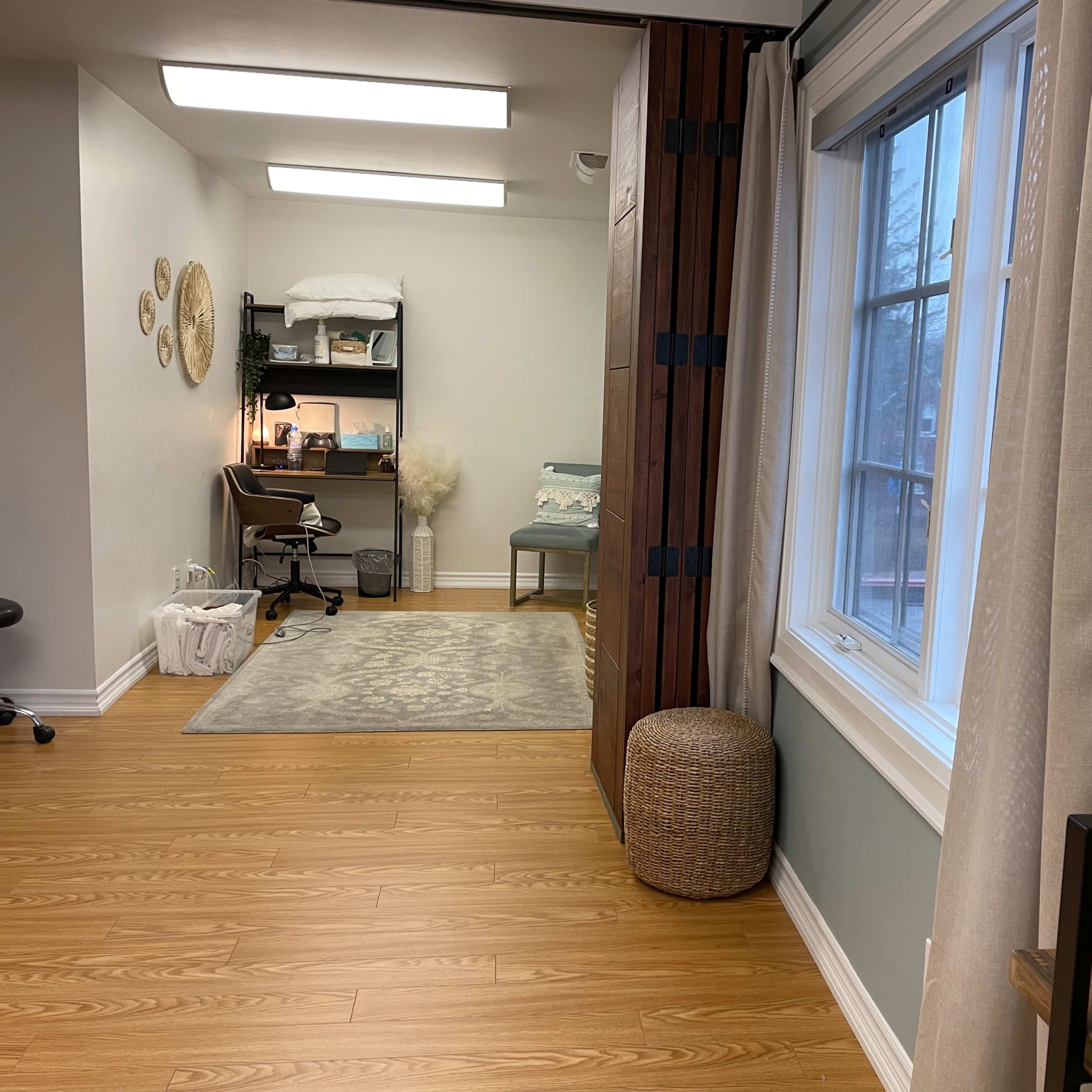 A well-lit room with wooden flooring, featuring a workspace with a chair and computer, a bed with linens, a closet area, and decorative wall elements.