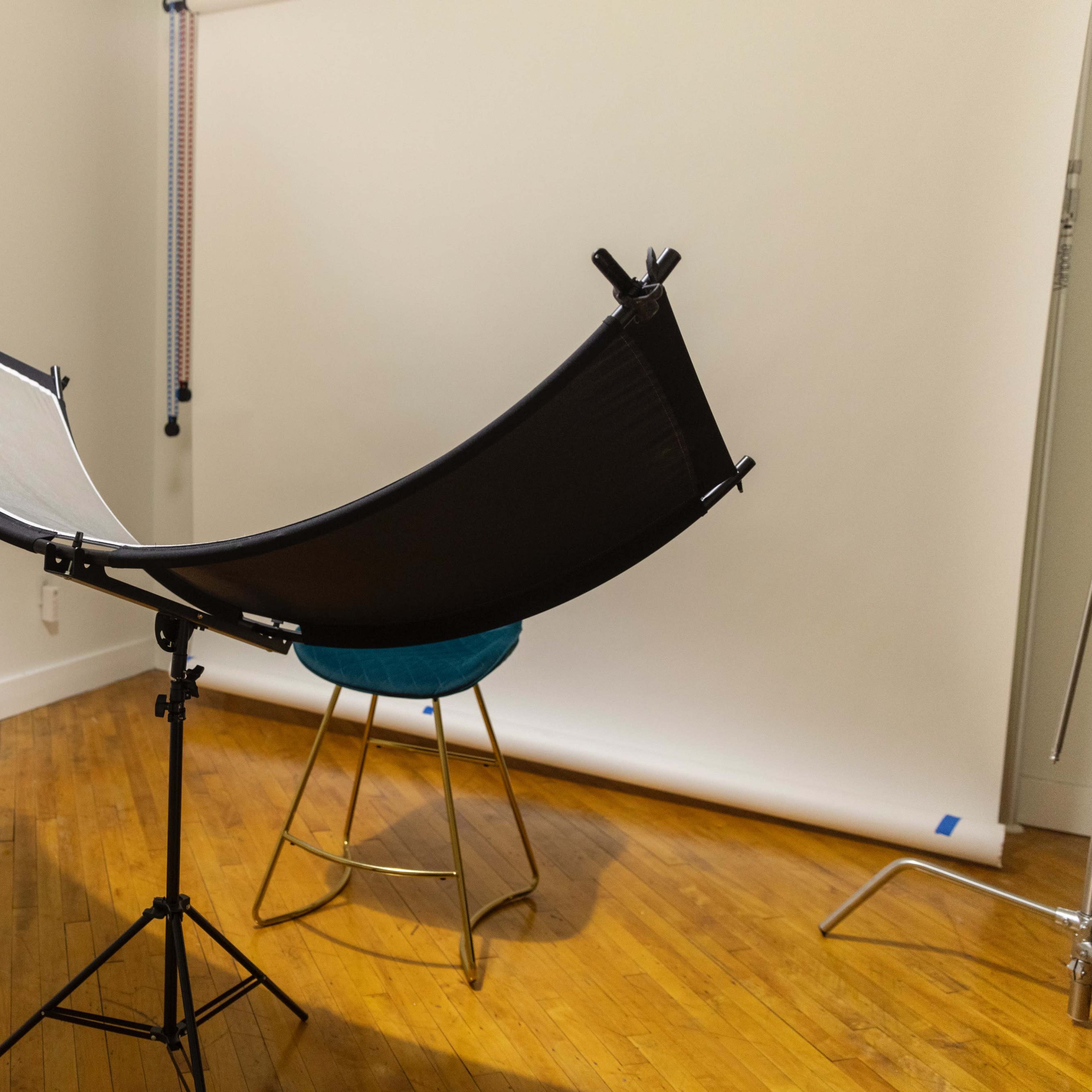 A black and white reflector is mounted on a tripod in an empty photography studio with wooden flooring and a stool nearby.