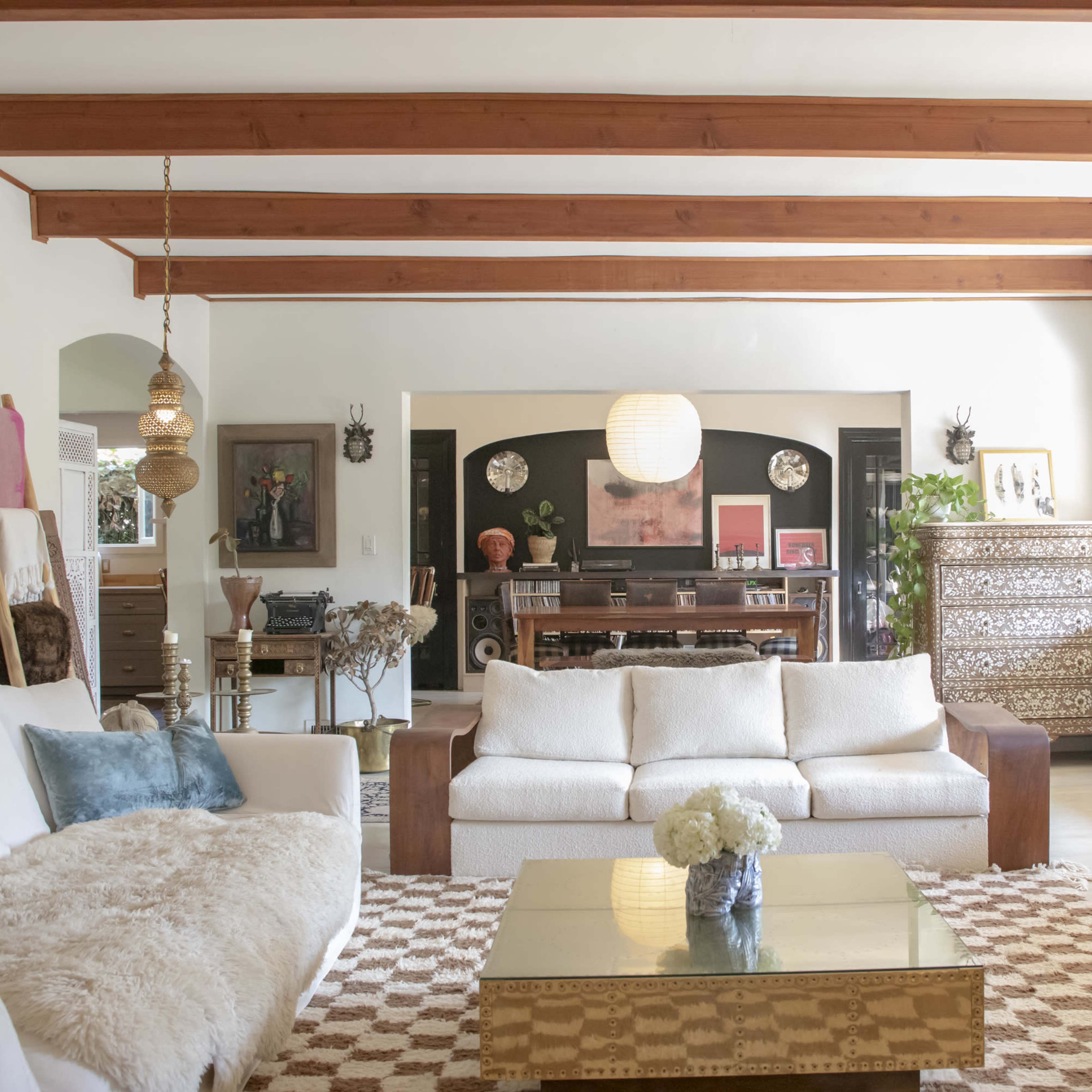A spacious living room features a light sofa, wooden beams above, a decorative coffee table, and a piano in the background.