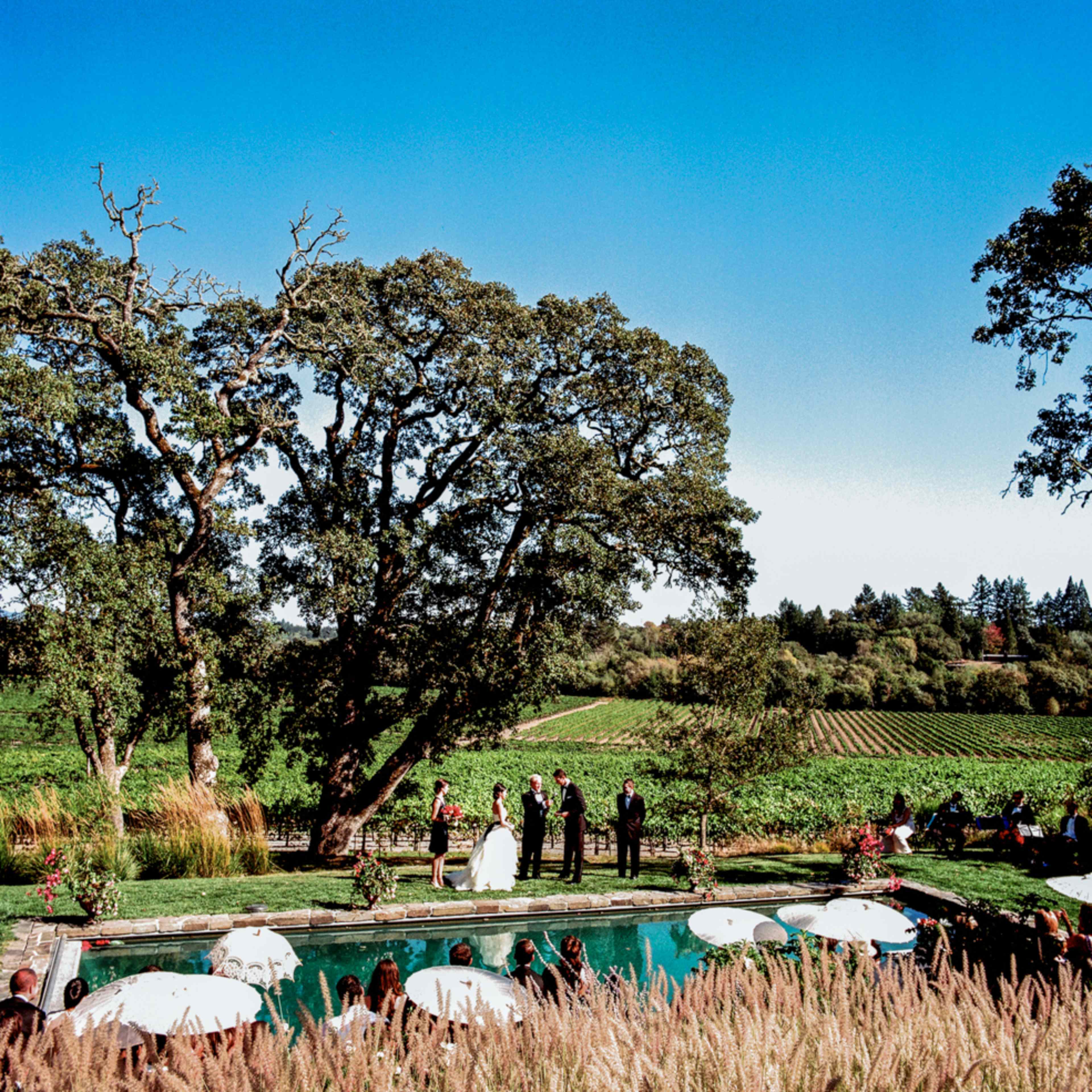 A wedding ceremony is taking place by a poolside in a vineyard, surrounded by large trees and guests seated under umbrellas.
