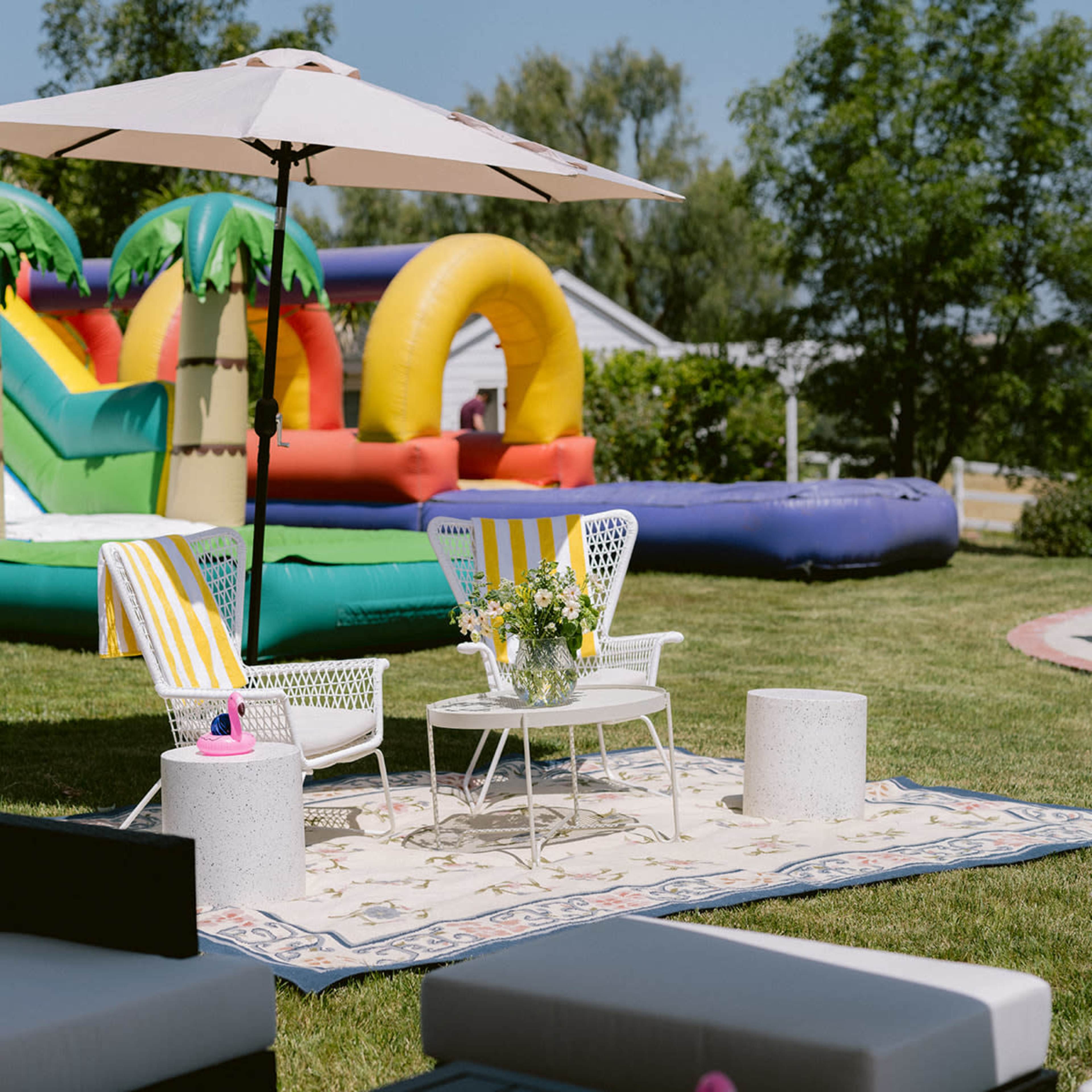 A colorful inflatable bounce house is set up in a grassy backyard, accompanied by a seating area featuring two white chairs and a small table.