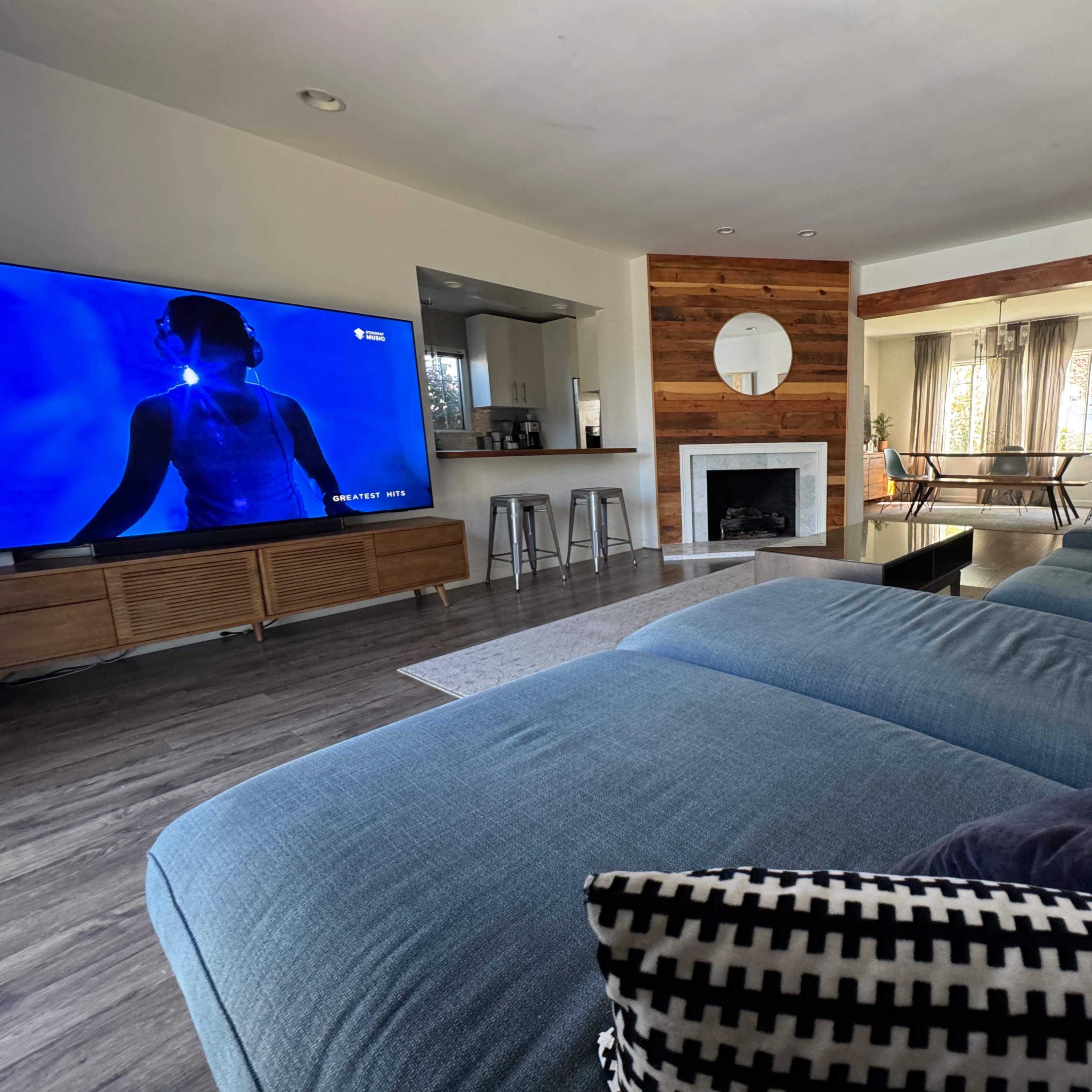 A modern living room featuring a large blue sectional sofa, a wall-mounted television displaying a figure, and an open kitchen area with a dining space in the background.
