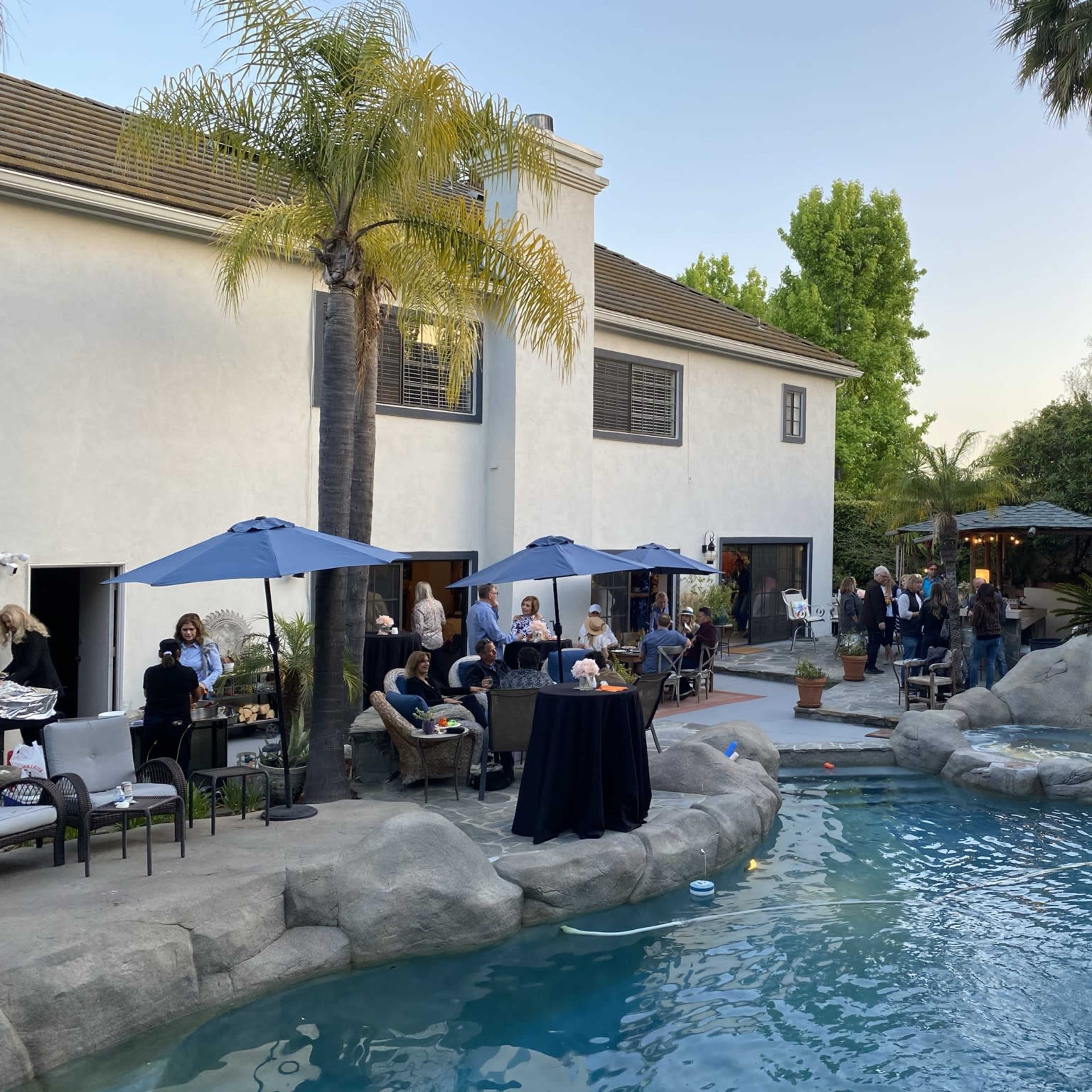 A gathering of people takes place in a backyard with a pool surrounded by rocks, palm trees, and a house in the background.
