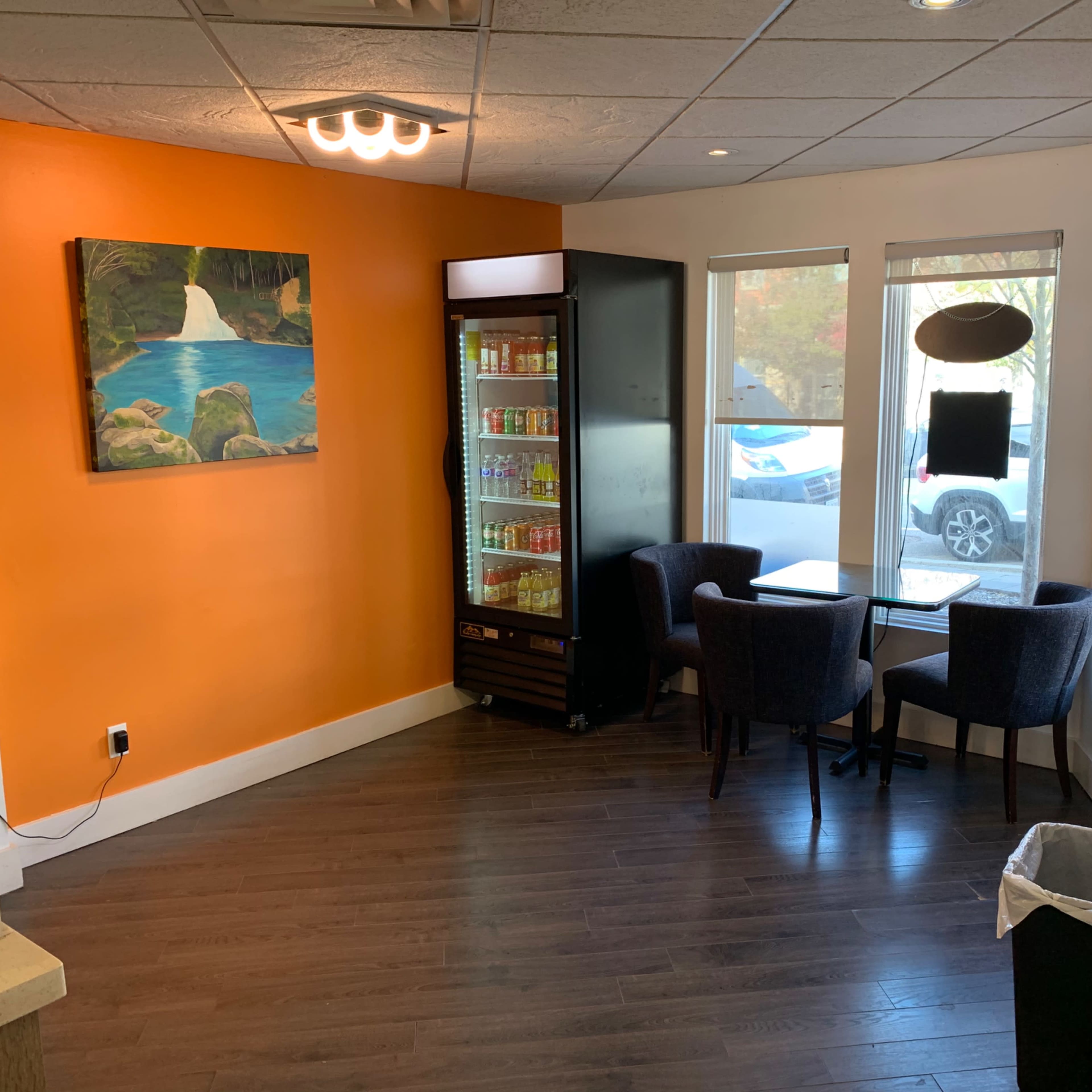 A small café corner with two chairs and a table beside a drink cooler, featuring an orange wall and a landscape painting.