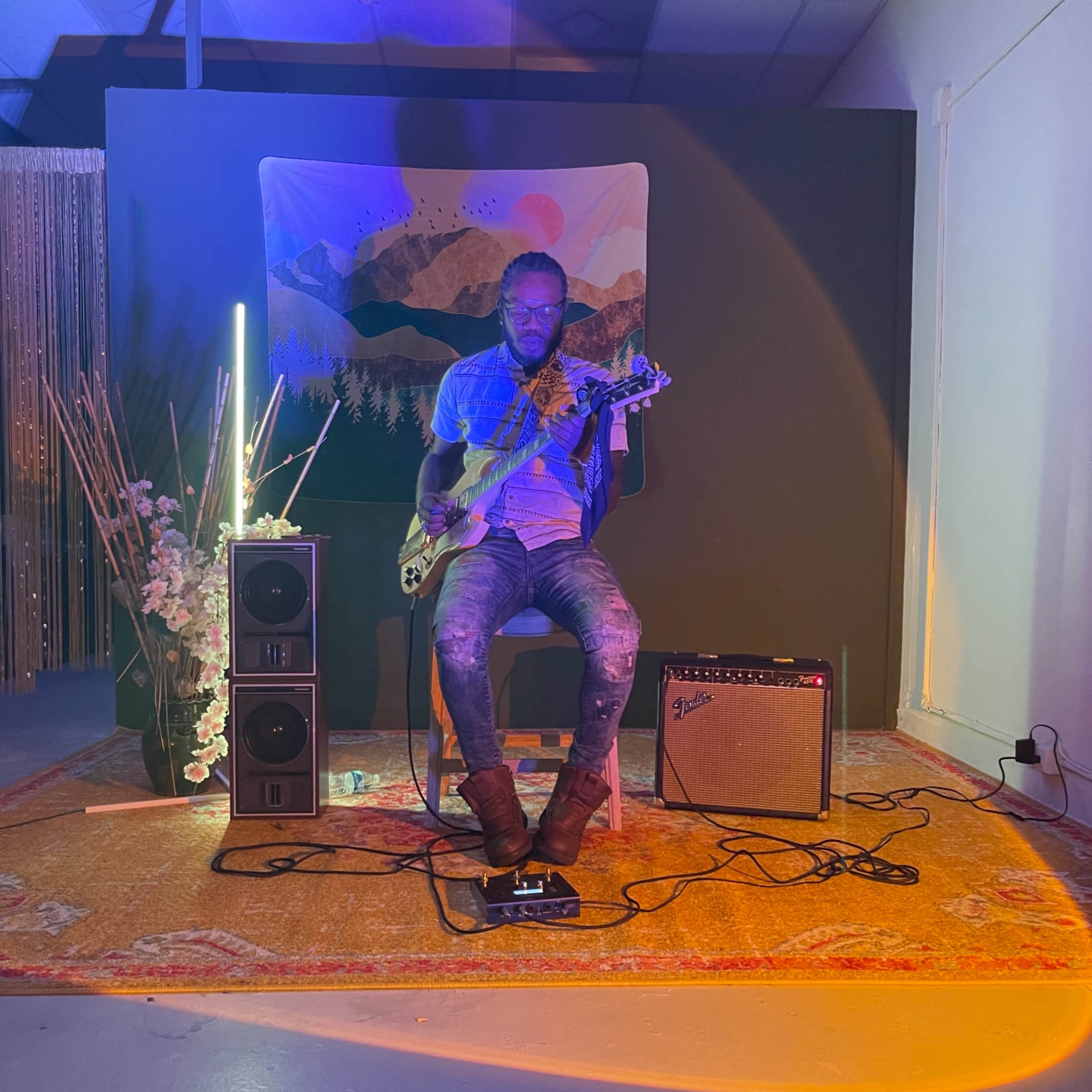 A musician plays an electric guitar while seated on a stool in a dimly lit room decorated with an art backdrop and musical equipment.