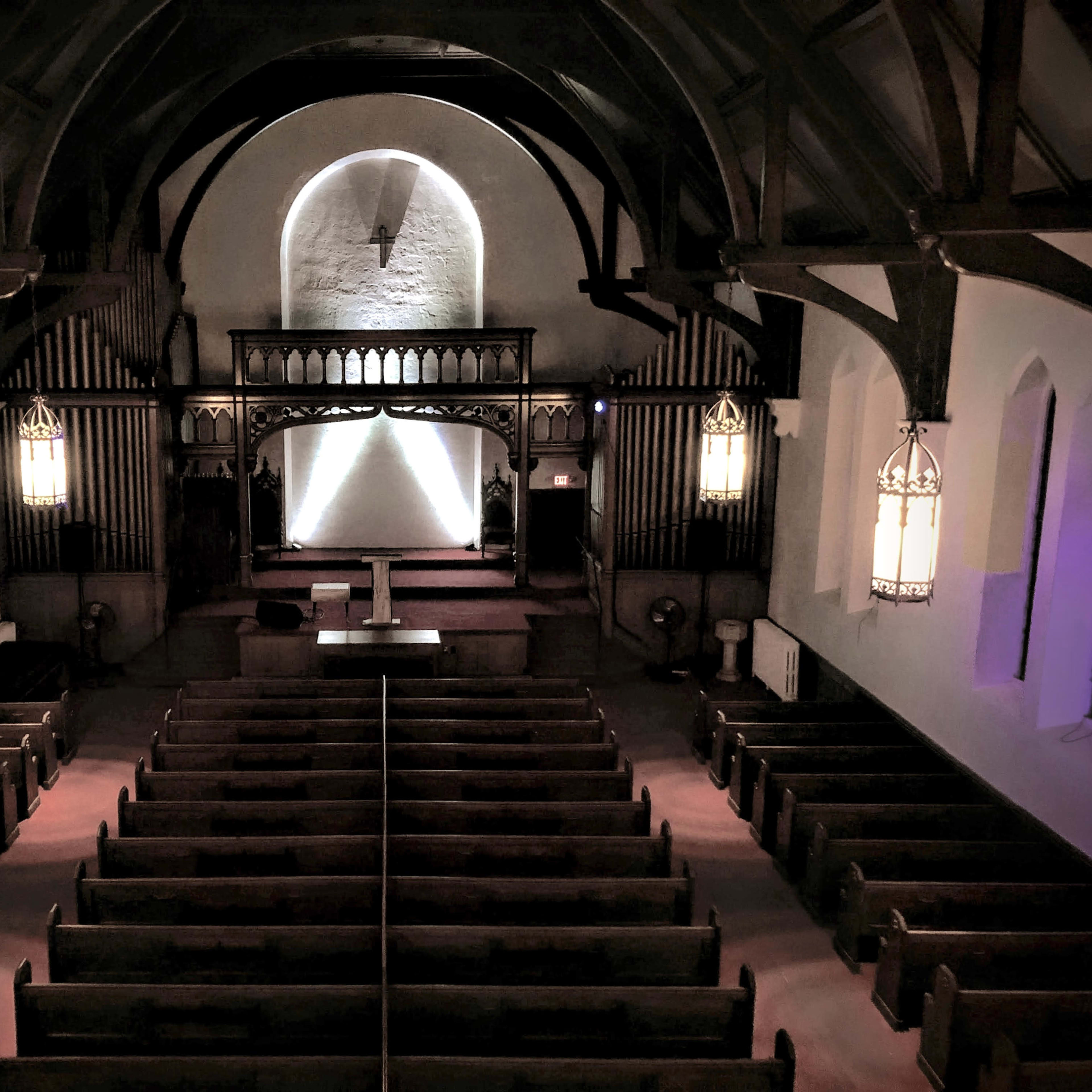 The interior of a church with wooden pews arranged in rows facing an altar at the front, illuminated by pendant lights.