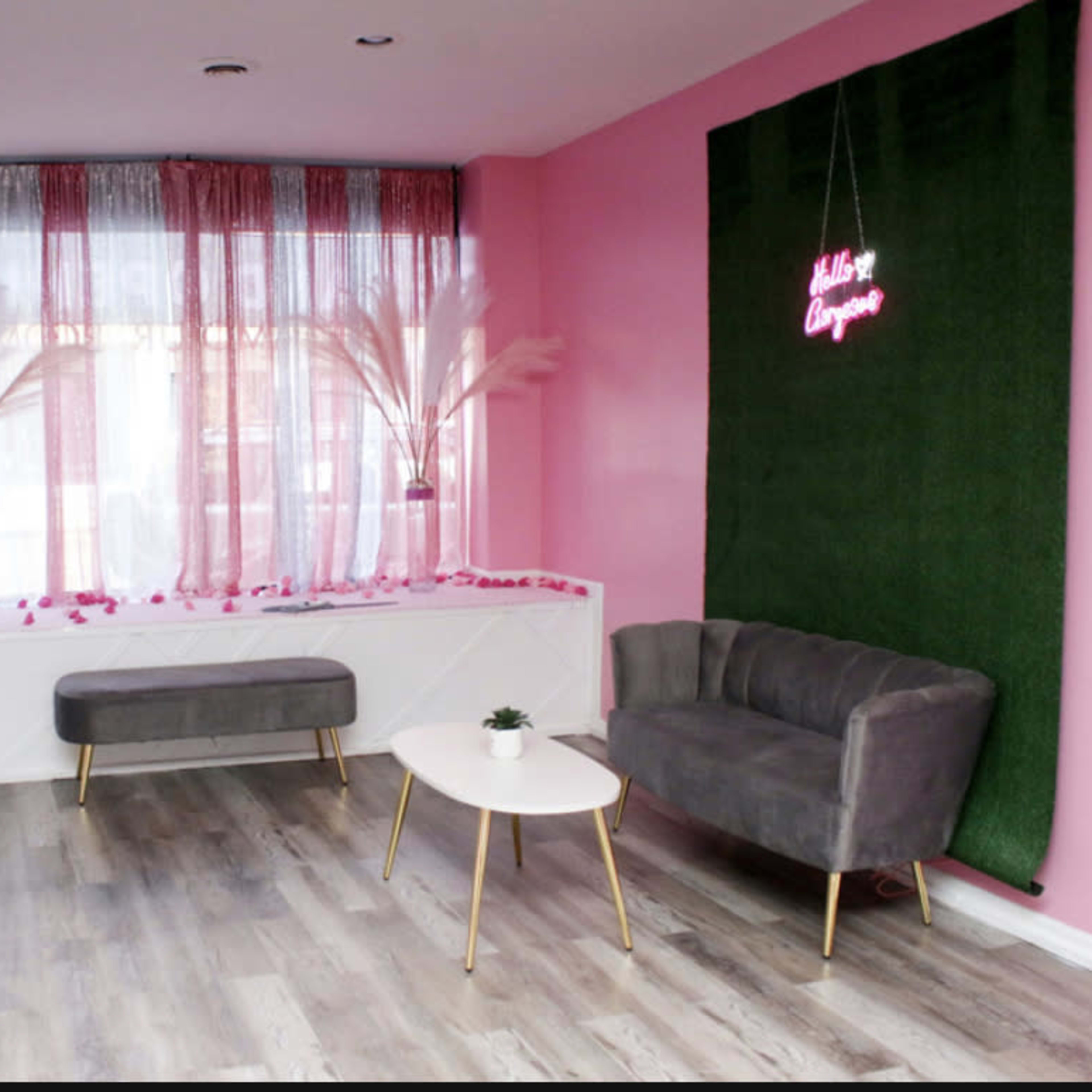 A modern indoor space with pink walls, a faux grass feature, a small table, and two grey sofas, accented by decorative plants and neon signage.