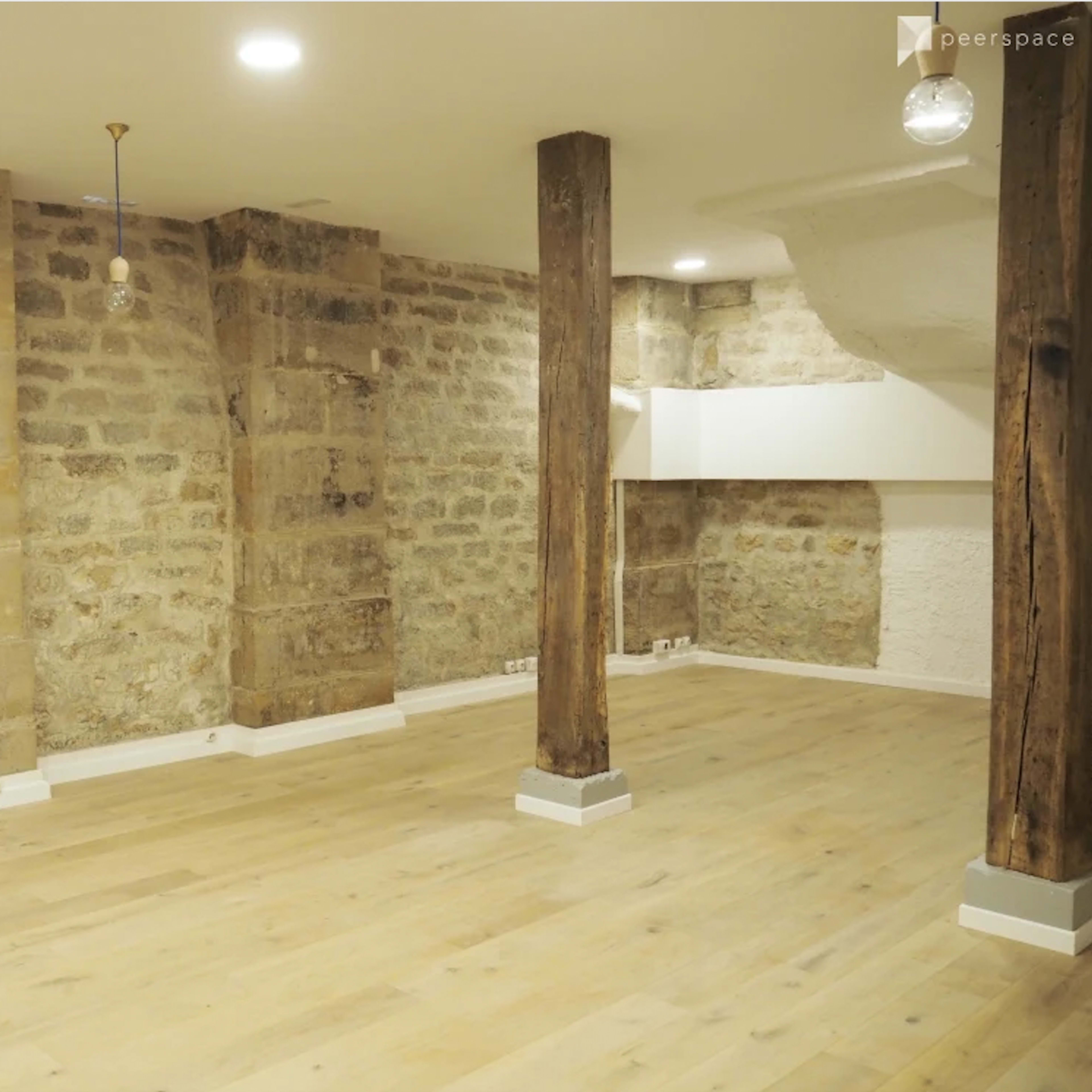 A spacious, empty interior room featuring stone walls, wooden pillars, and wooden flooring, with modern pendant lights hanging from the ceiling.
