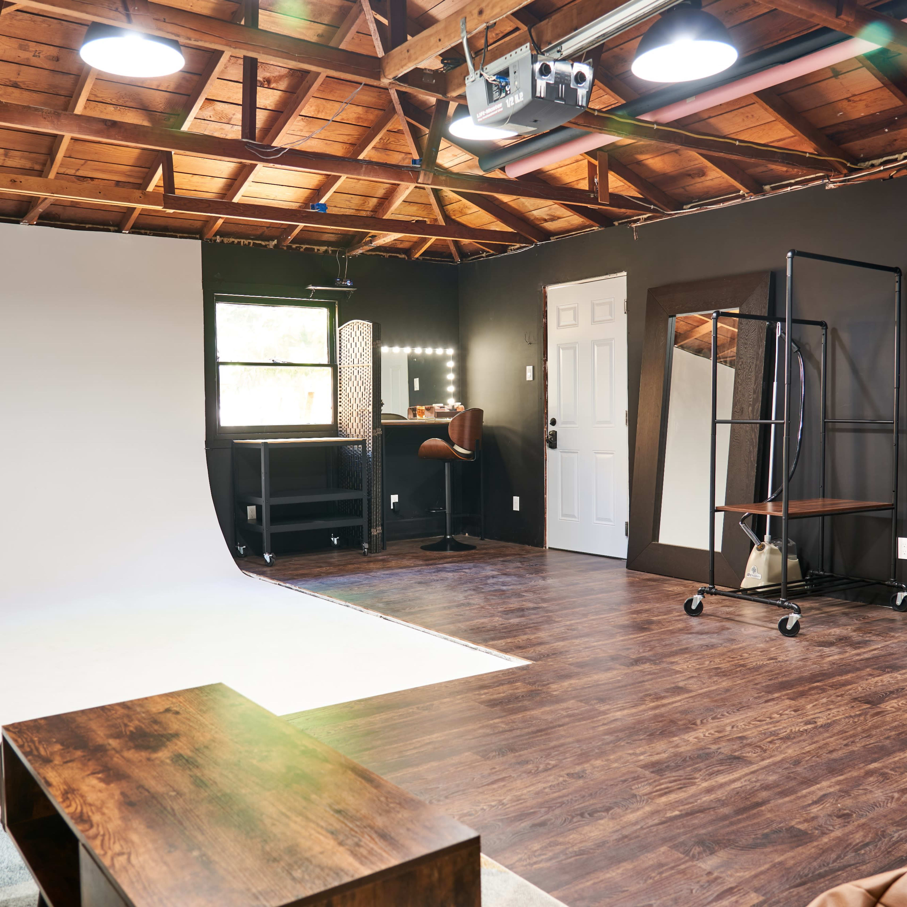 The interior of a studio features a white backdrop, a makeup station with lights, a mirror, and a wooden coffee table on a dark wood floor.