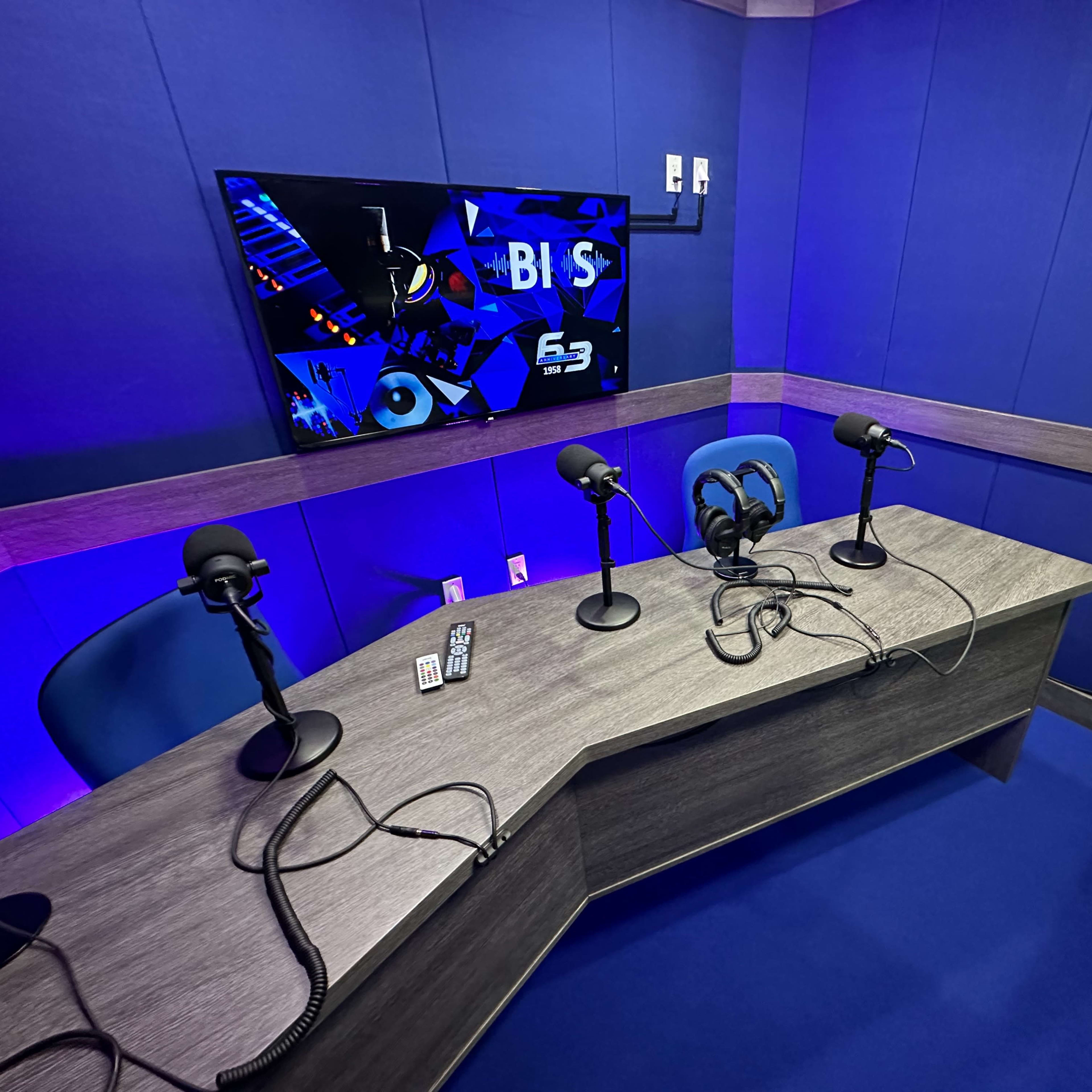 A modern recording studio with a blue backdrop, featuring a wooden desk and three microphones on stands.