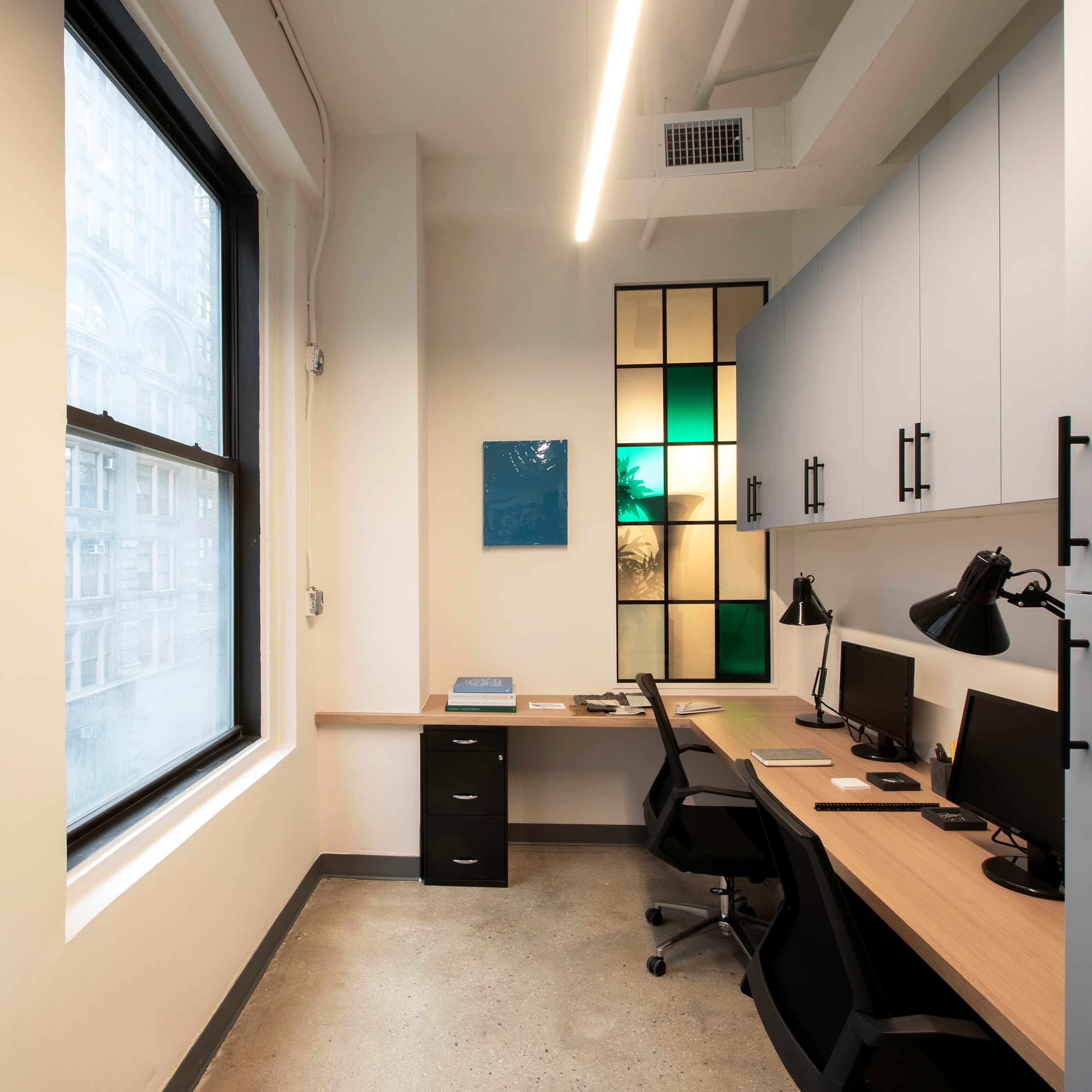 A modern office space with two desks, each equipped with a computer, beside large windows and a colorful glass panel.