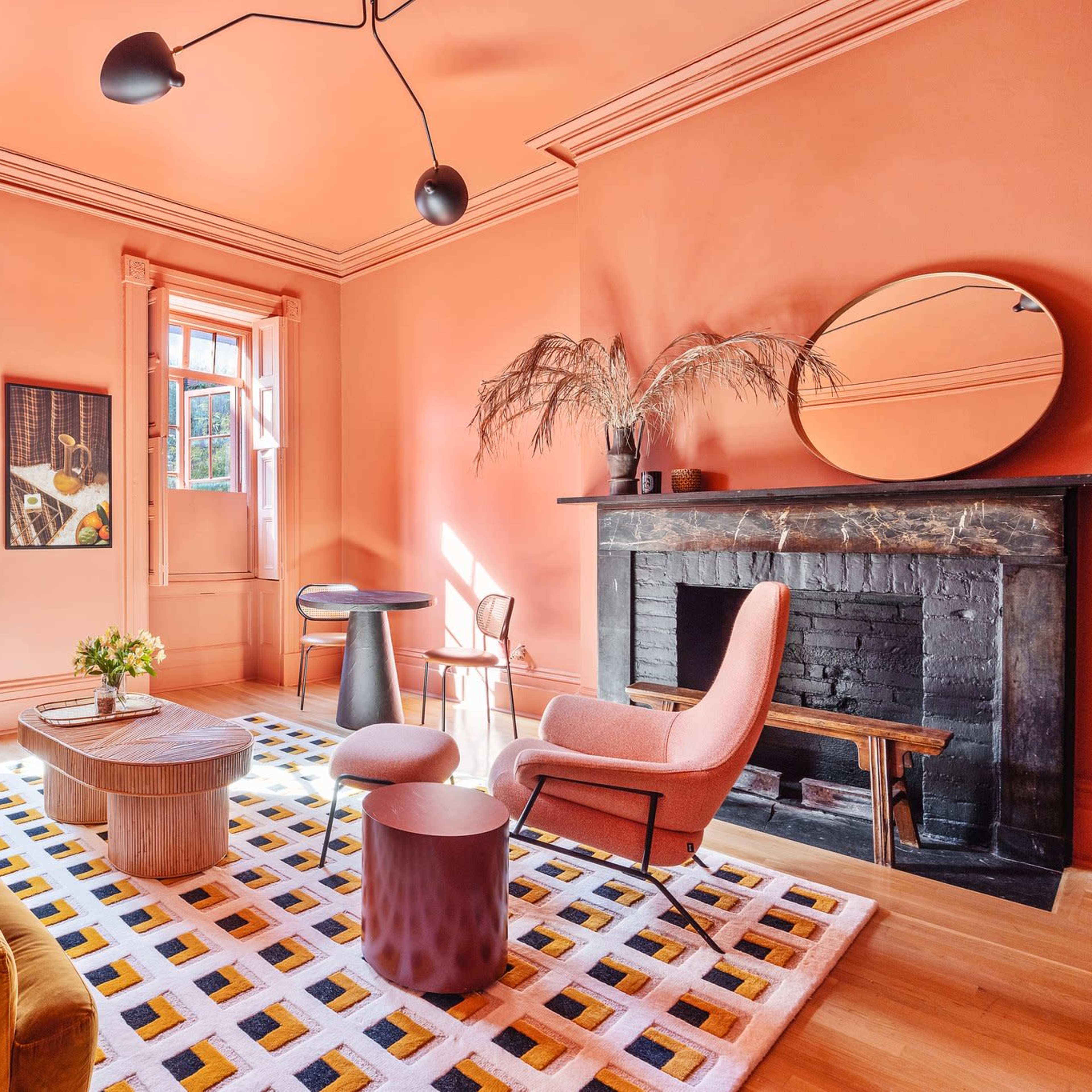 A well-lit living room featuring a peach-colored wall, a yellow sofa, a round wooden coffee table, and a black fireplace, with large windows allowing natural light to enter.