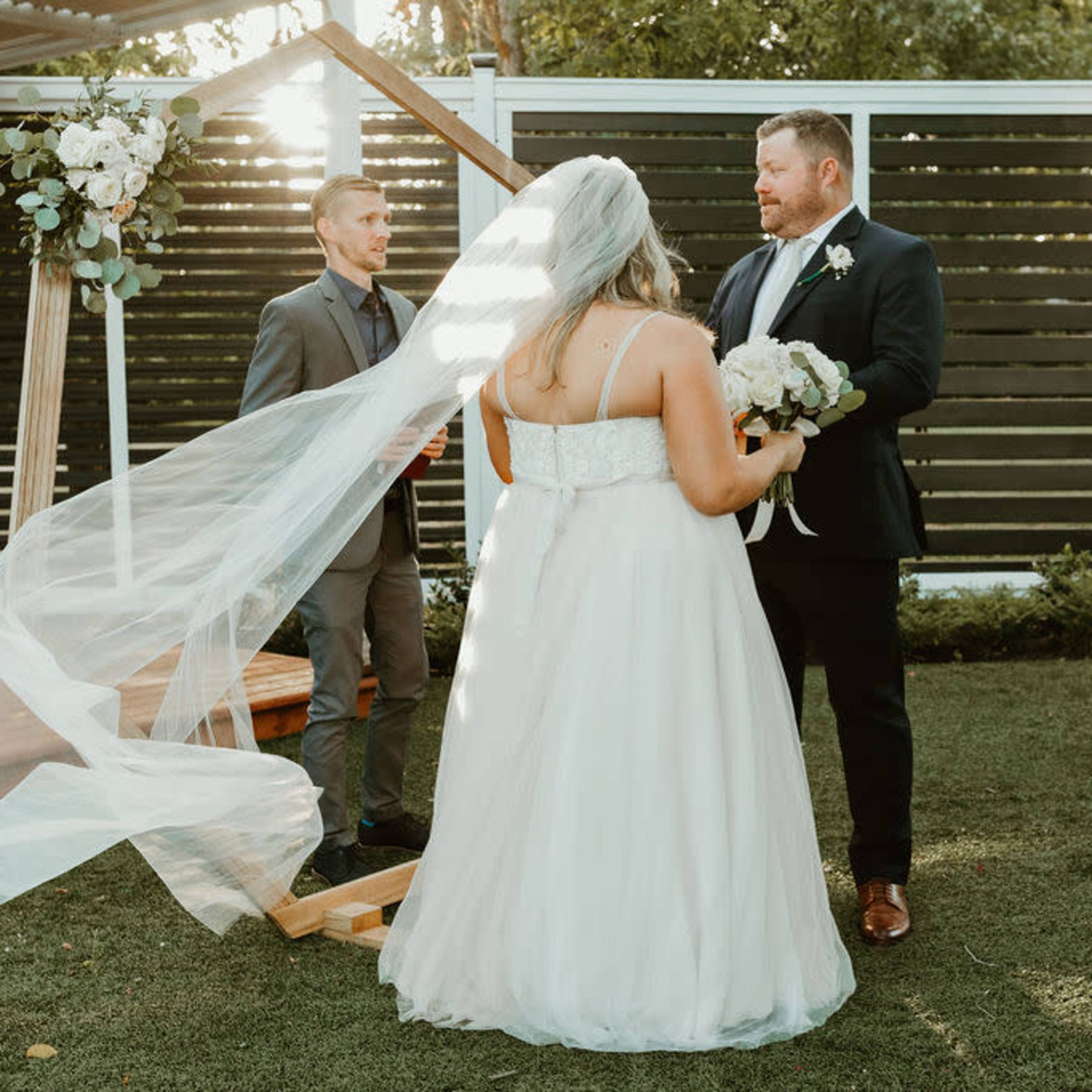 A bride in a white gown with a long veil stands in front of a groom during a wedding ceremony under a wooden structure in a garden setting.