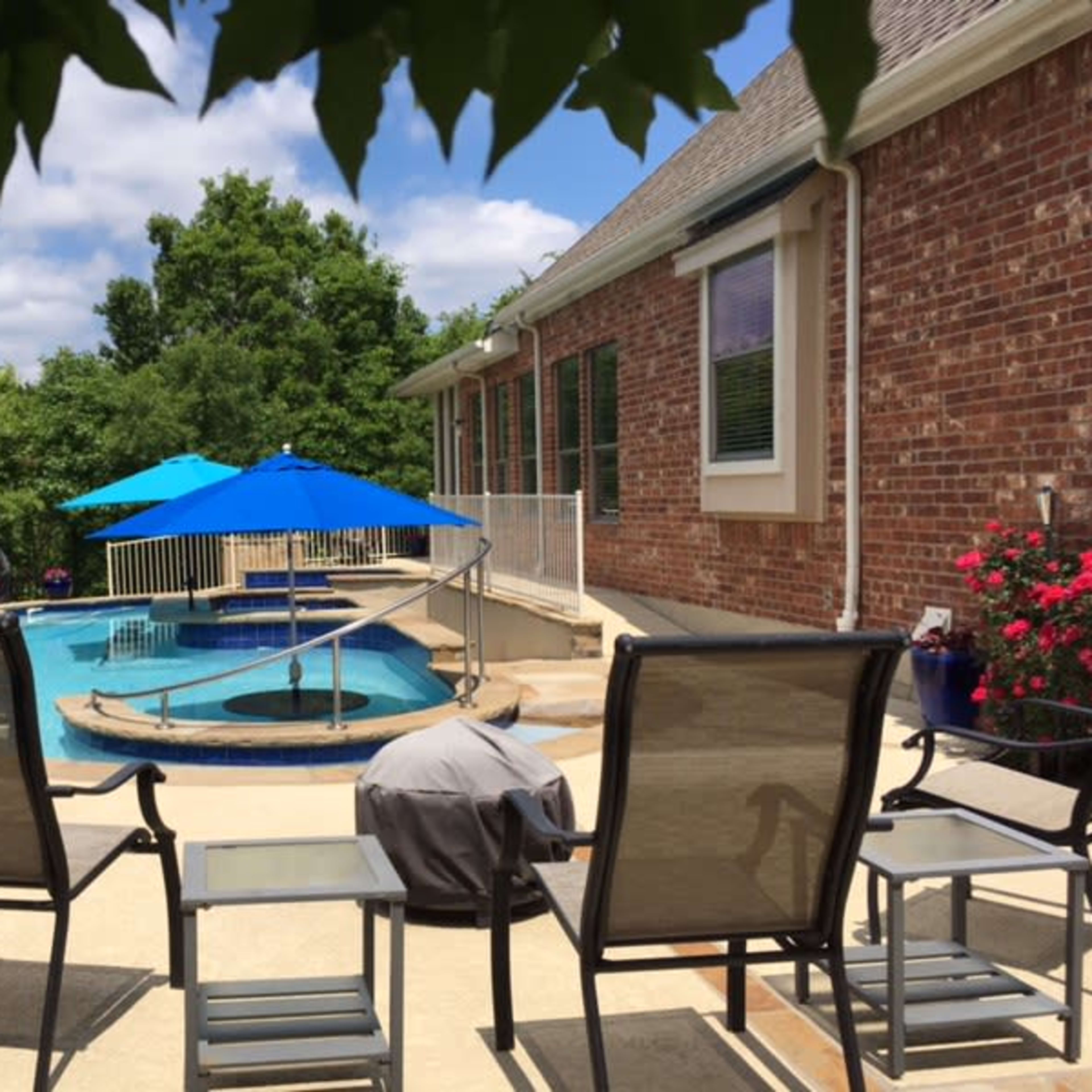 A patio features several chairs facing a blue pool surrounded by greenery and a brick house.
