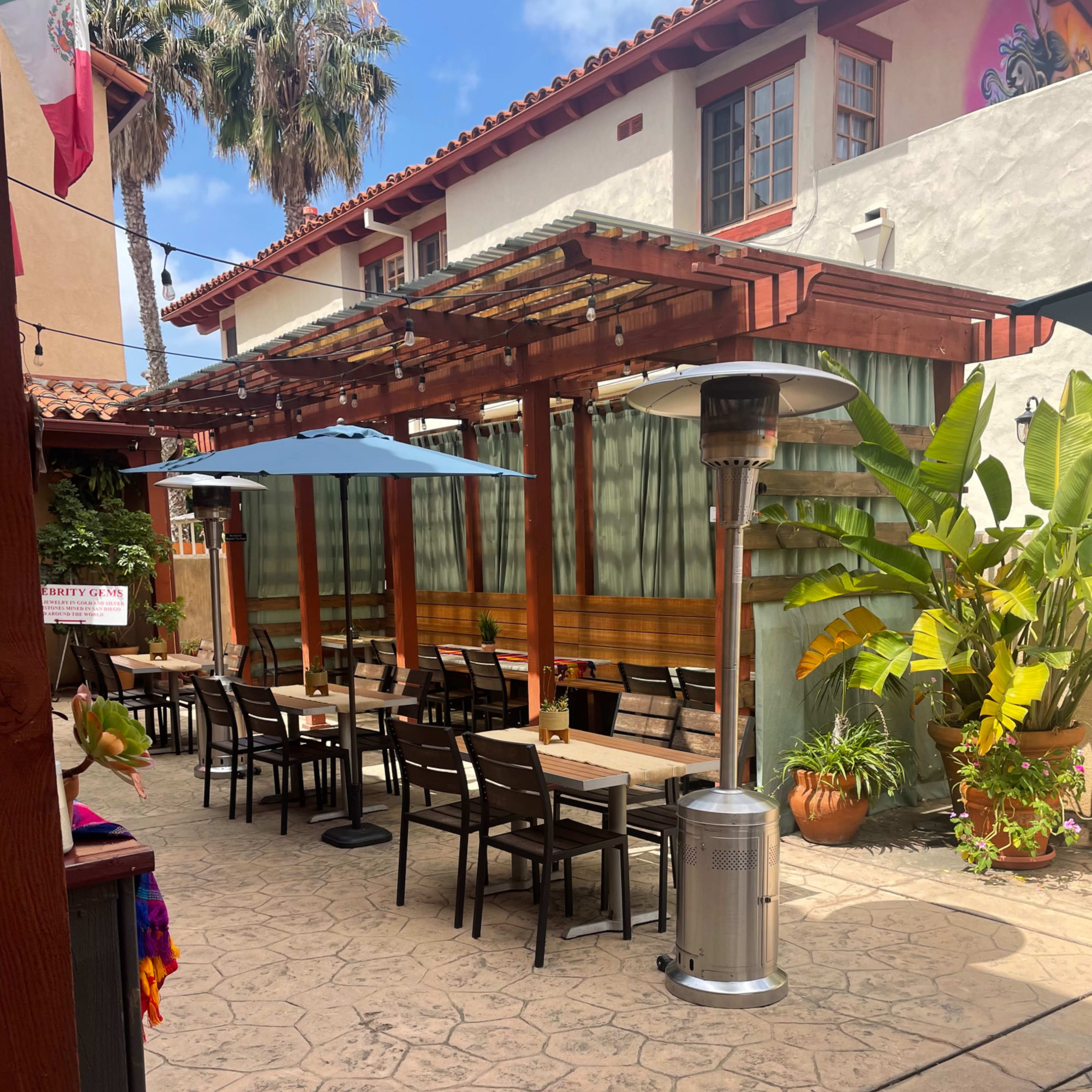A sunlit outdoor dining area features wooden tables and chairs, shaded by umbrellas and surrounded by tropical plants.