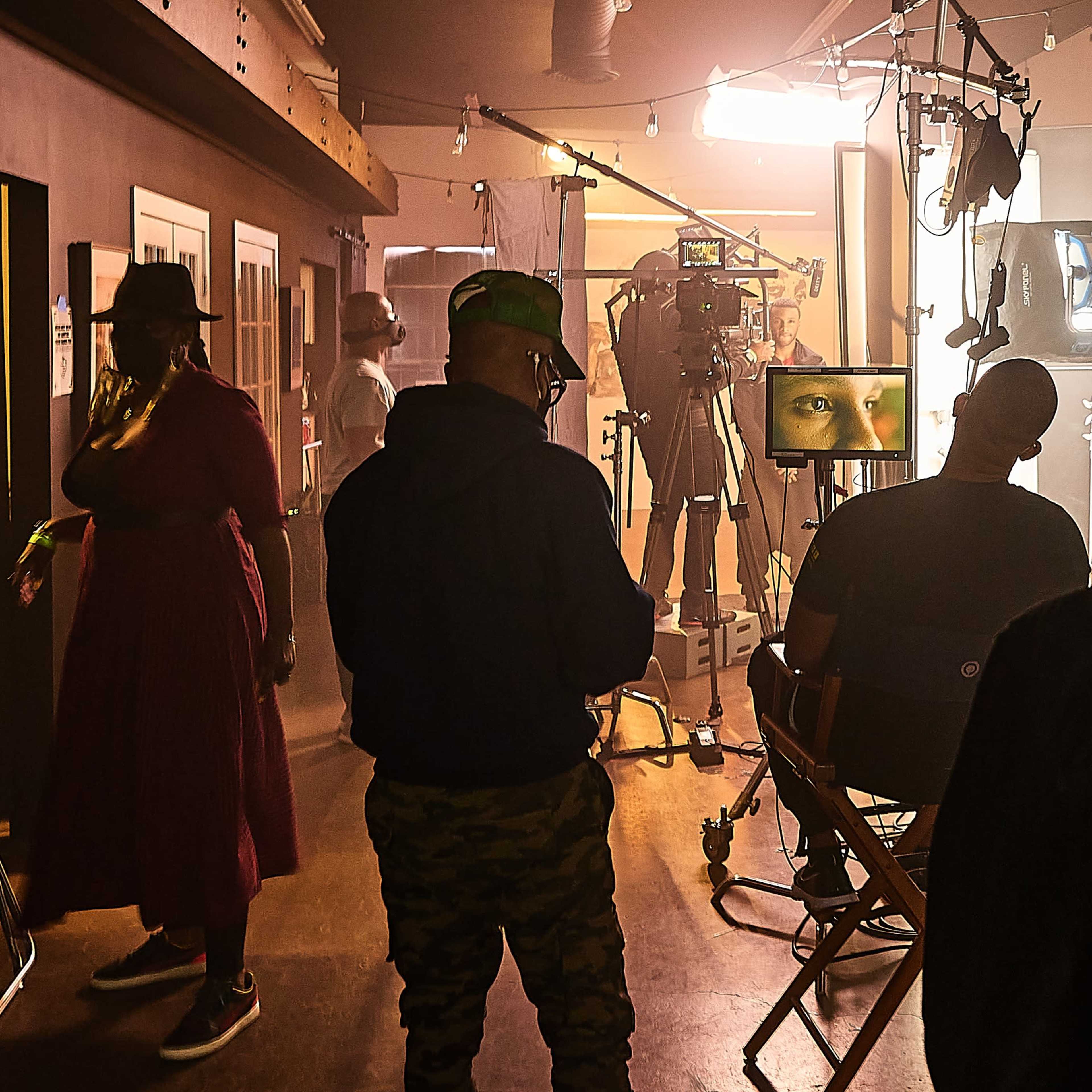 A group of people is gathered in a studio setting, observing a filming setup with cameras and lighting equipment focused on a seated individual.