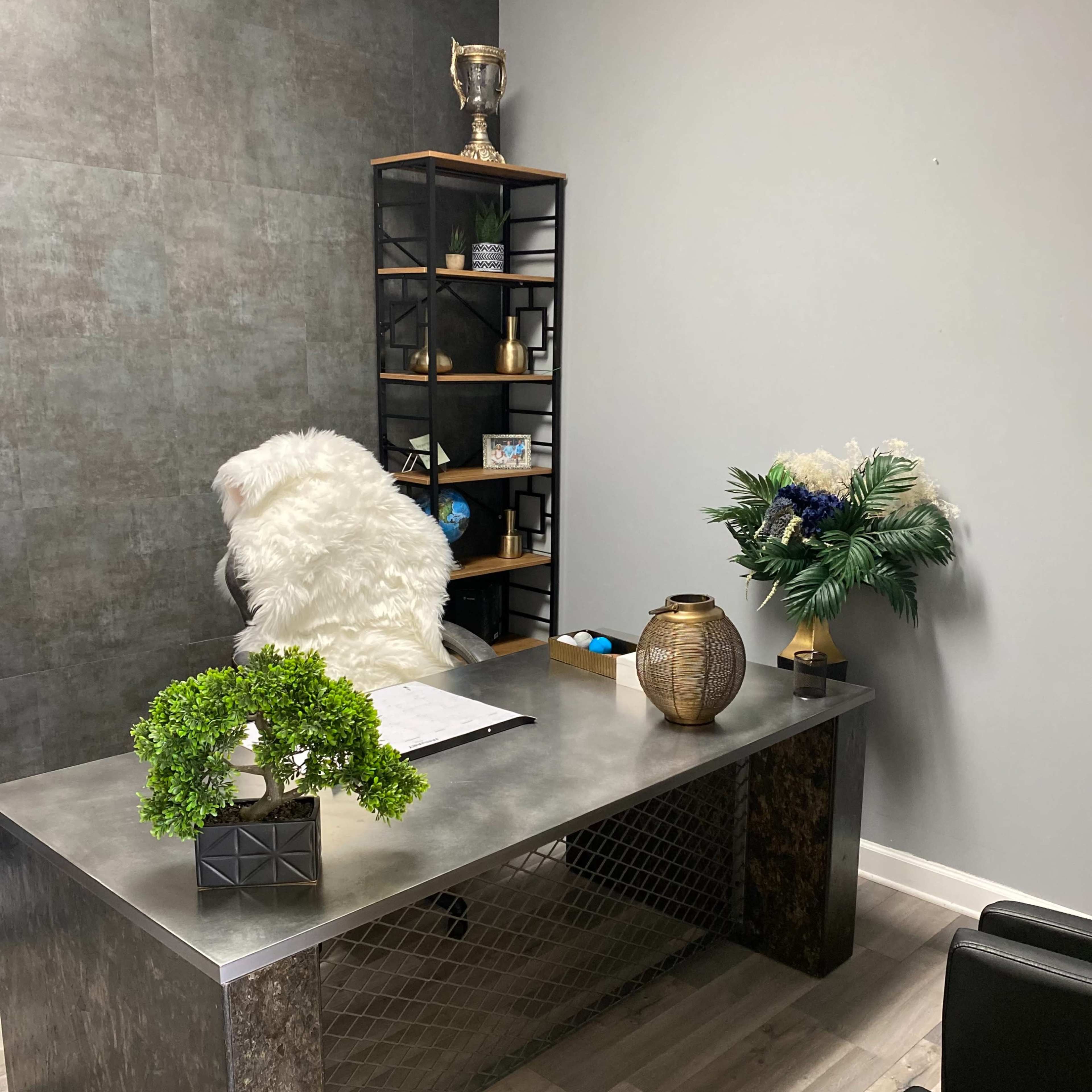A modern office space with a metal desk, a plush white chair, a bookshelf with decorative items, and a small arrangement of plants and flowers.