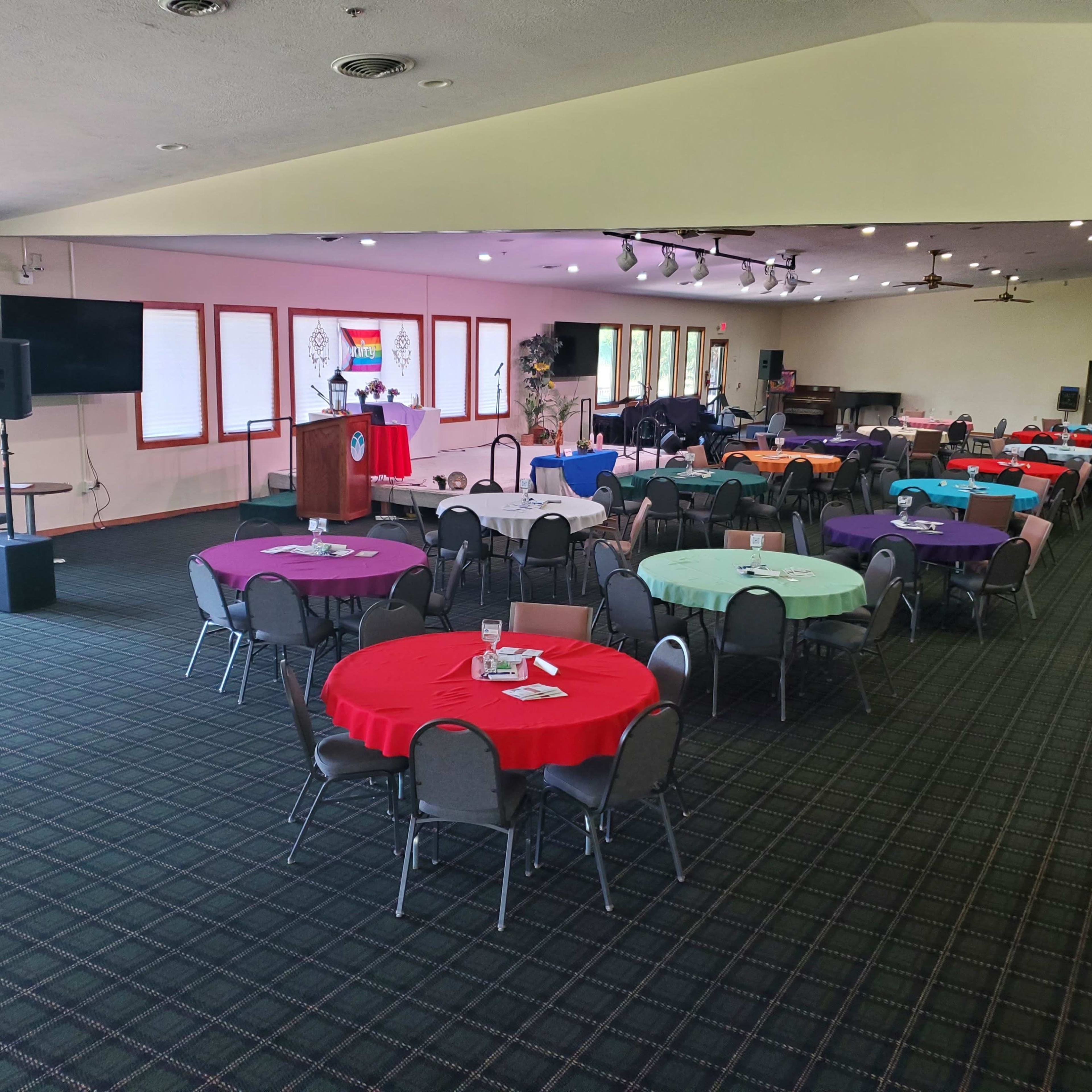 A spacious event room features multiple tables covered in colorful tablecloths arranged in rows, with a podium and a stage in the background.
