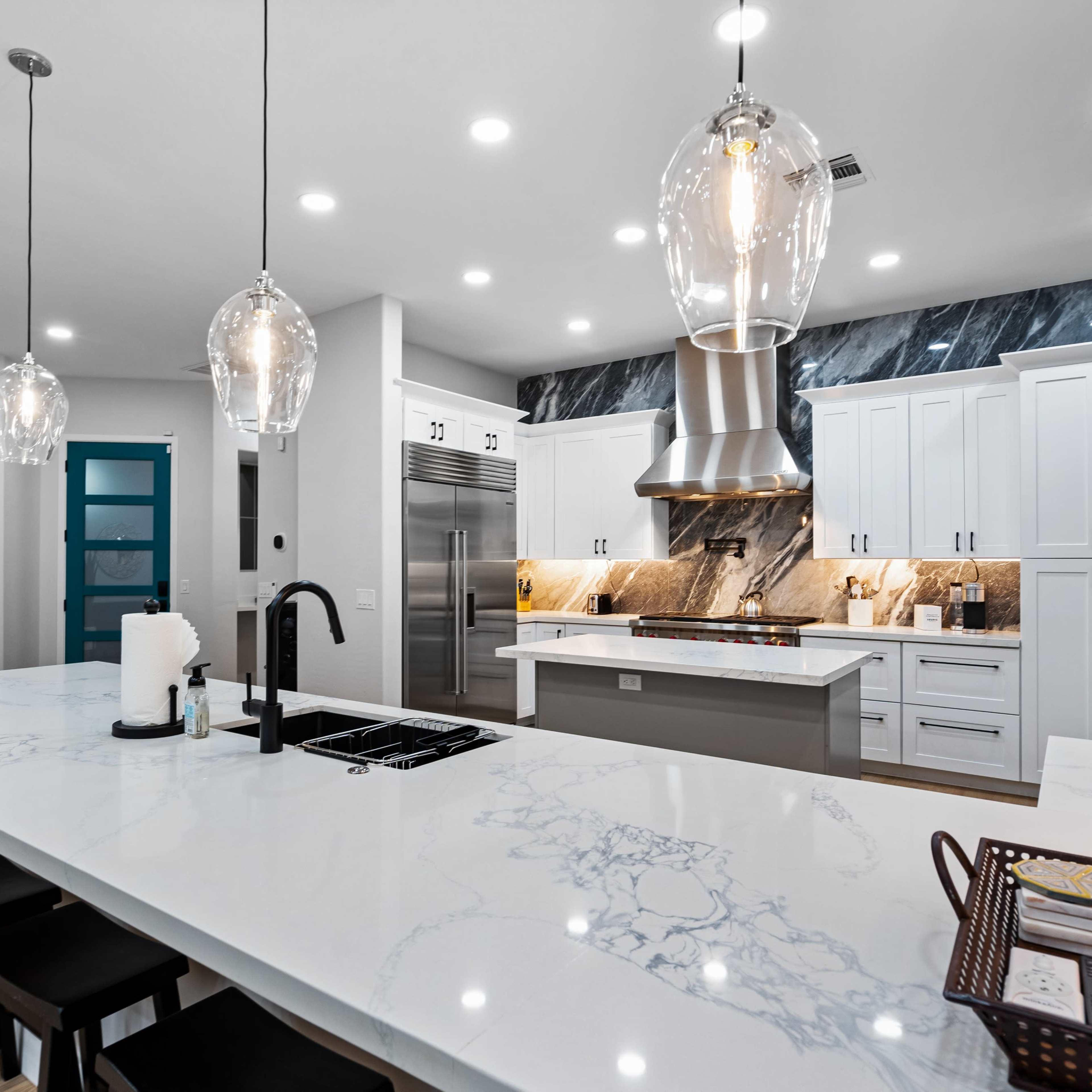 A modern kitchen features a large island with a marble countertop, pendant lighting, and stainless steel appliances.
