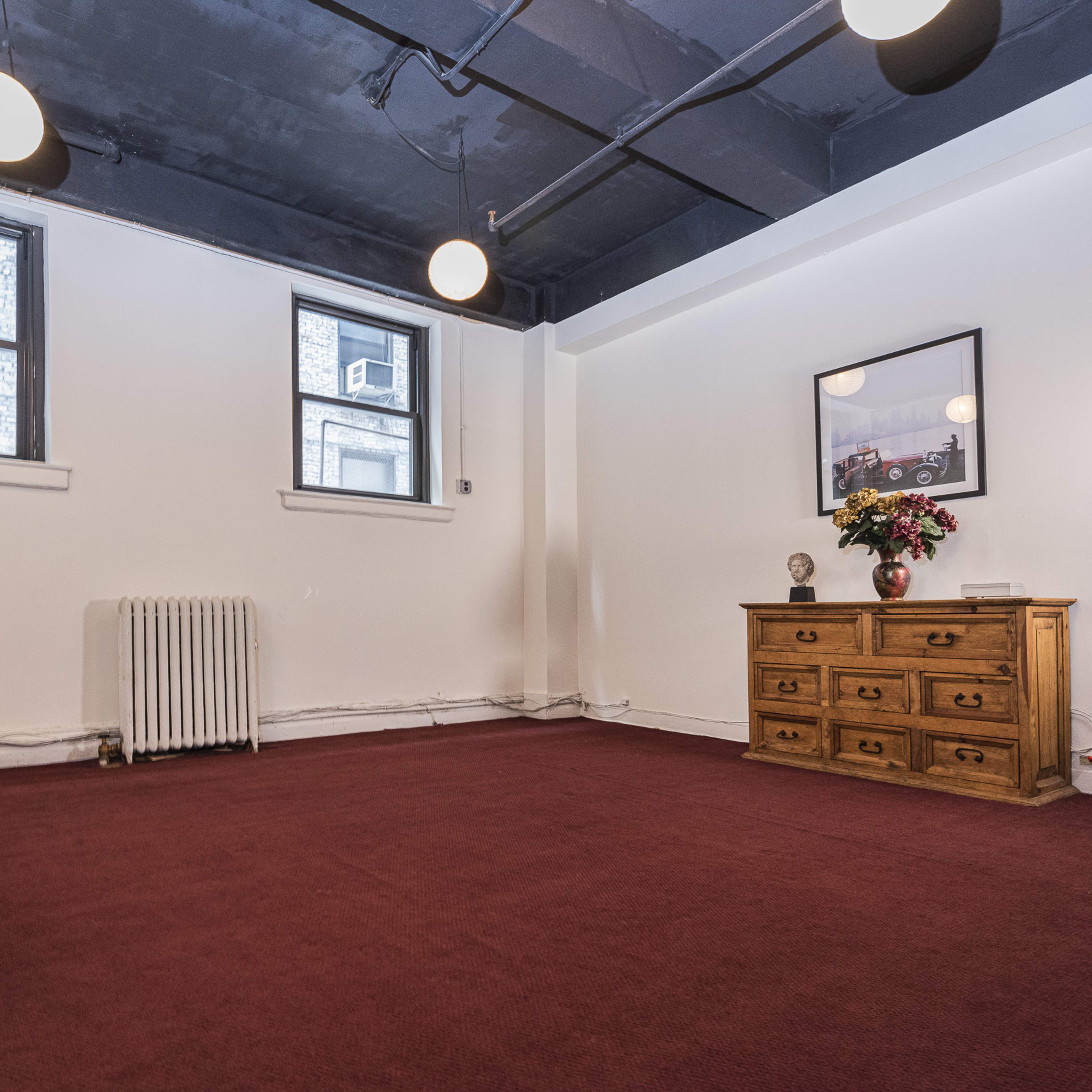 A spacious room with carpeted flooring, two windows, and a wooden cabinet with drawers and a vase of flowers on a side table.