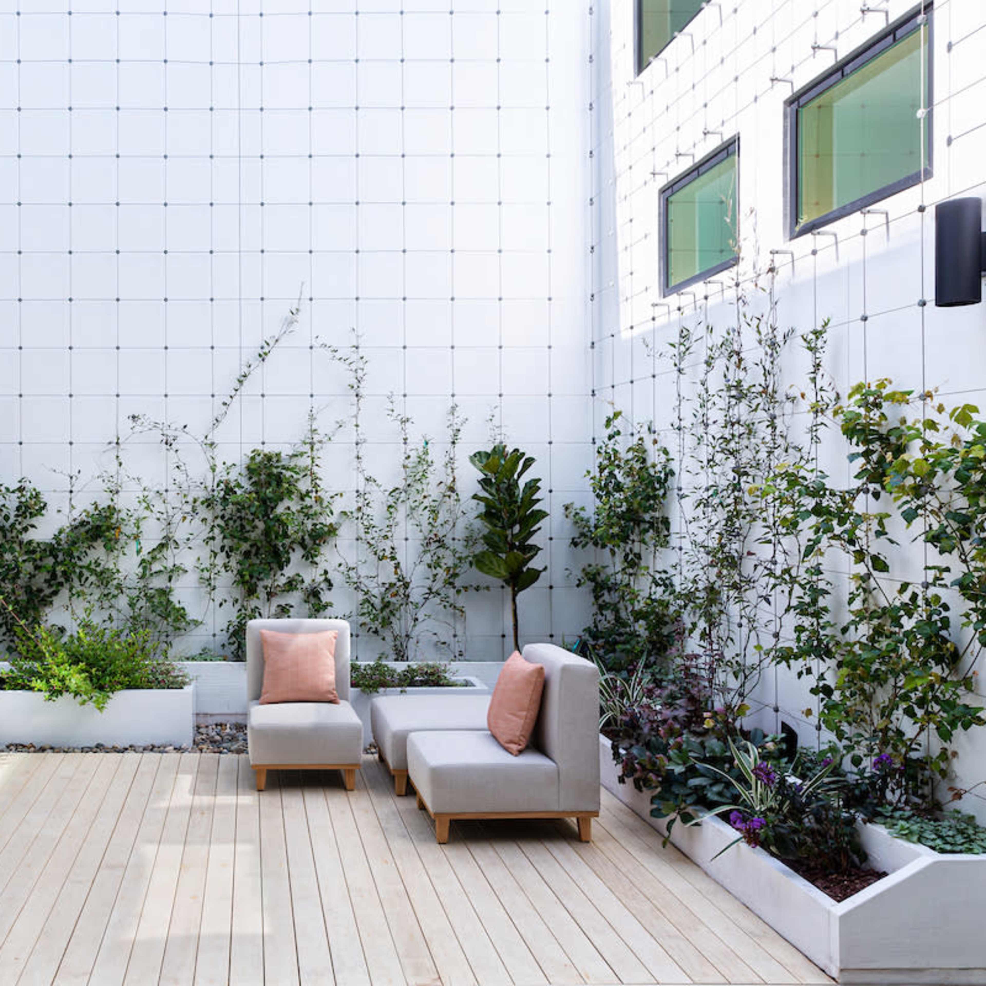 A minimalist outdoor space features a wooden deck with two gray lounge chairs surrounded by plants in white planters against a tiled wall.