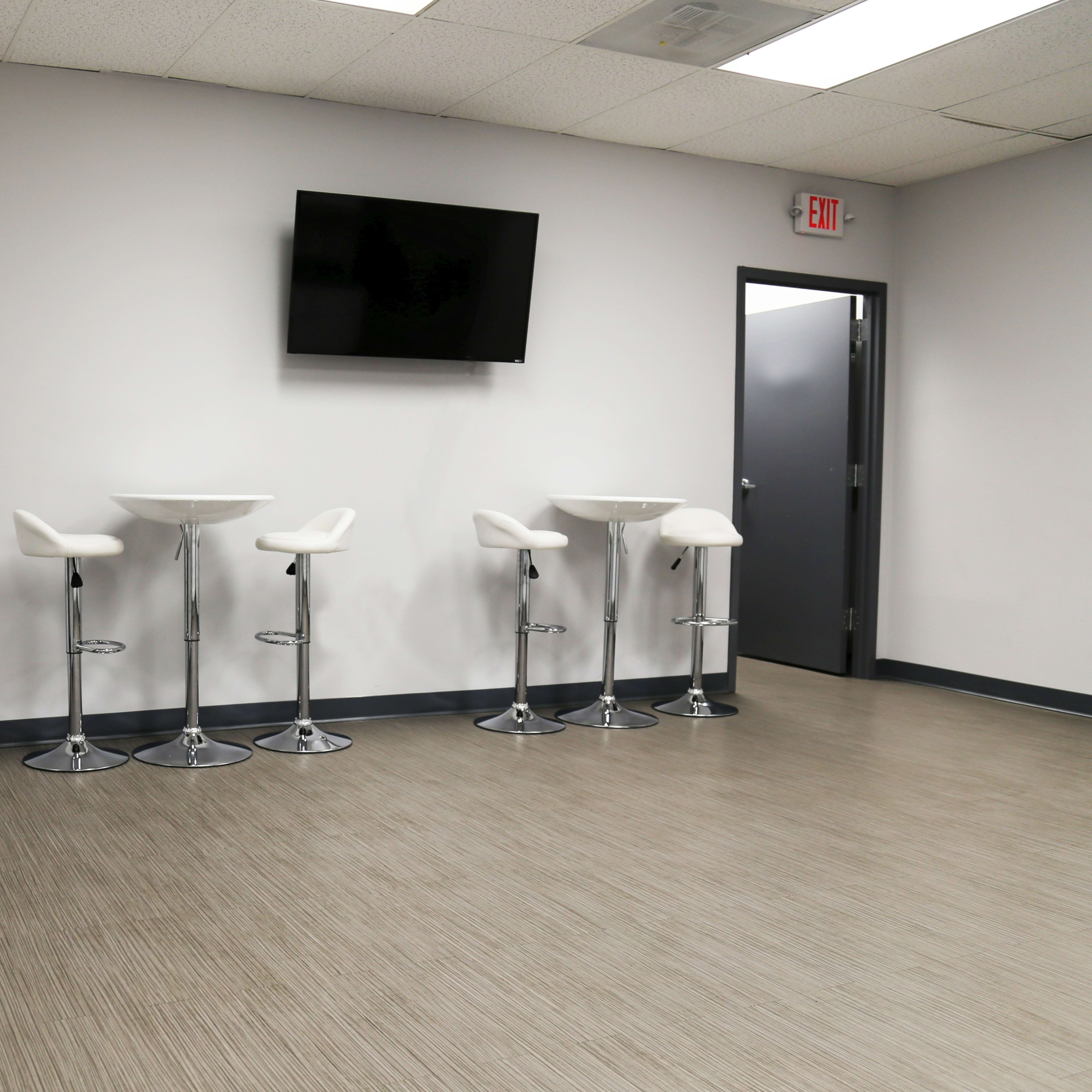 A plain room with four high stools and small tables against a light gray wall, featuring a wall-mounted television.