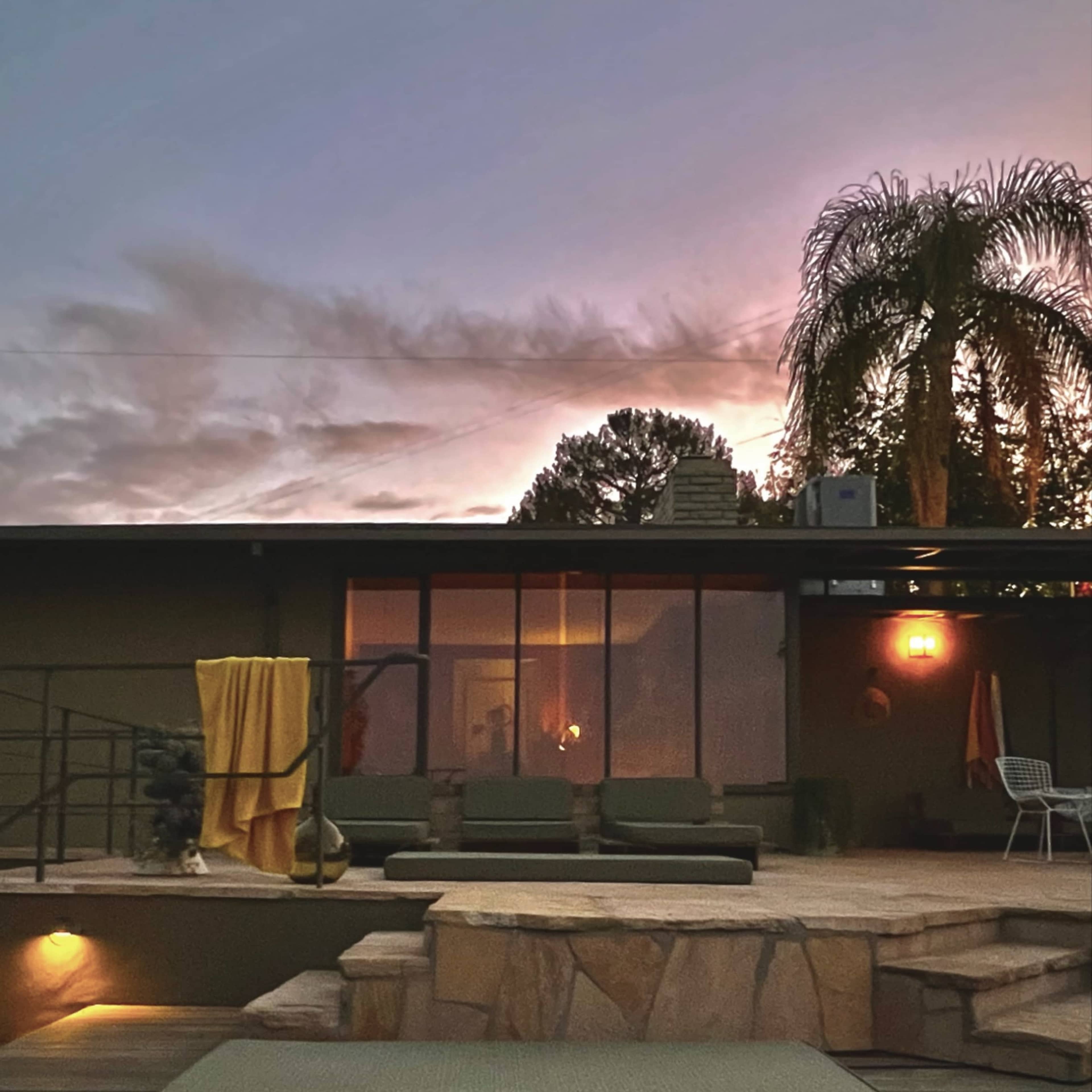 A modern home features a stone patio and pool, with outdoor seating and a warm glow in the windows as the sun sets.