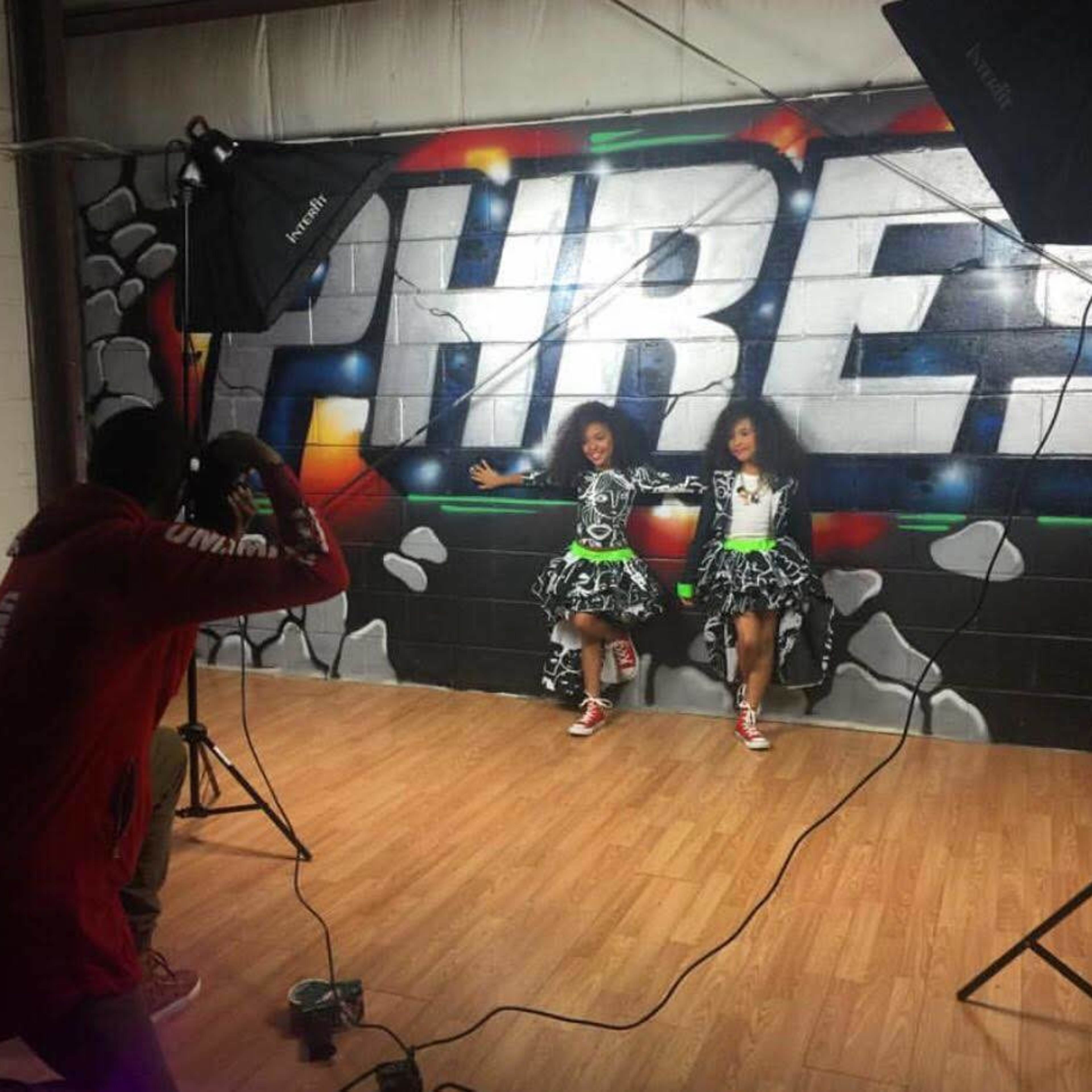 Two dancers pose in colorful outfits against a graffiti-covered wall while a photographer captures the moment in a dance studio.