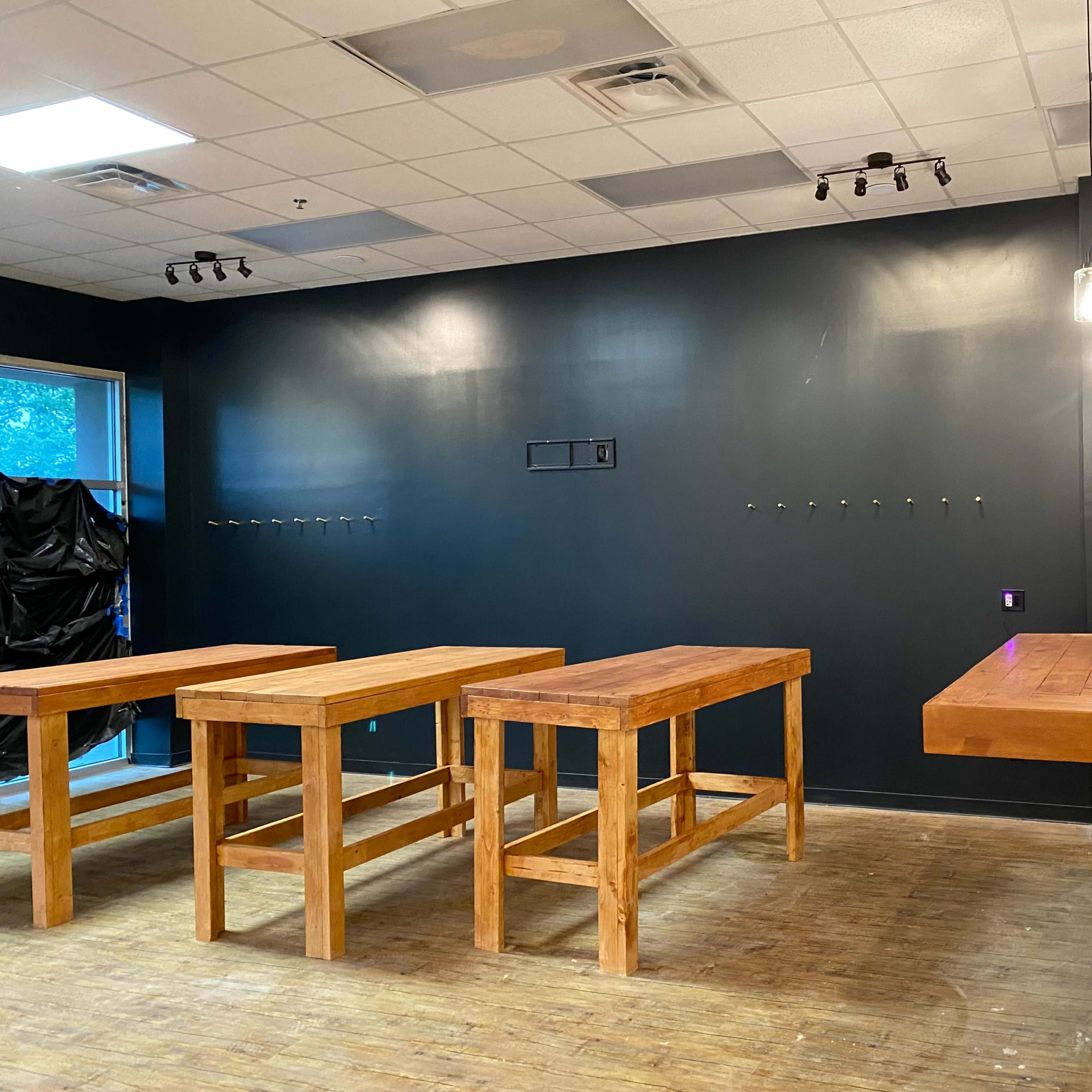 An interior space with several wooden tables arranged in a row and a dark wall with hooks, illuminated by overhead lighting.