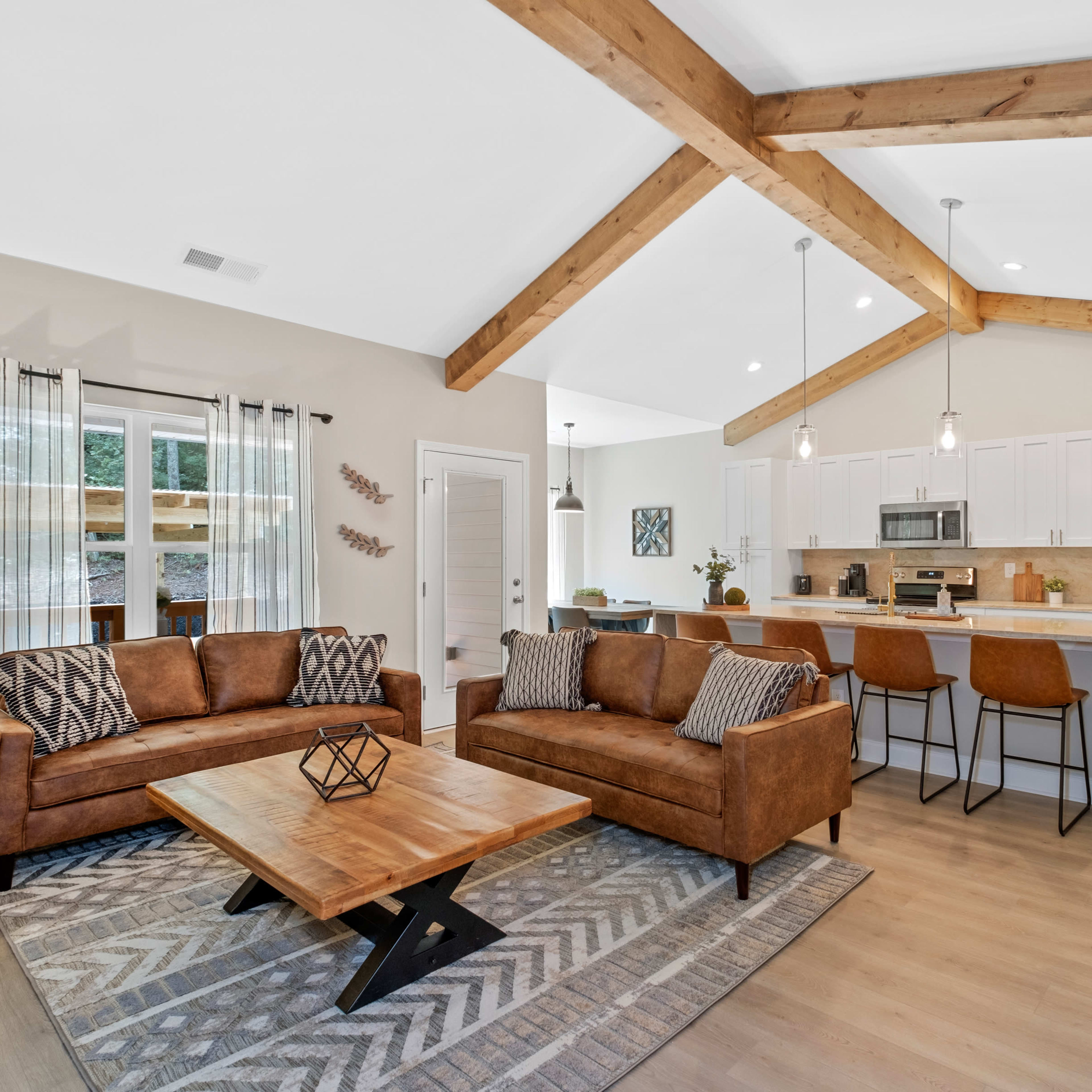 A modern living room with two brown sofas, a wooden coffee table, and a kitchen area in the background featuring white cabinets and stainless steel appliances.