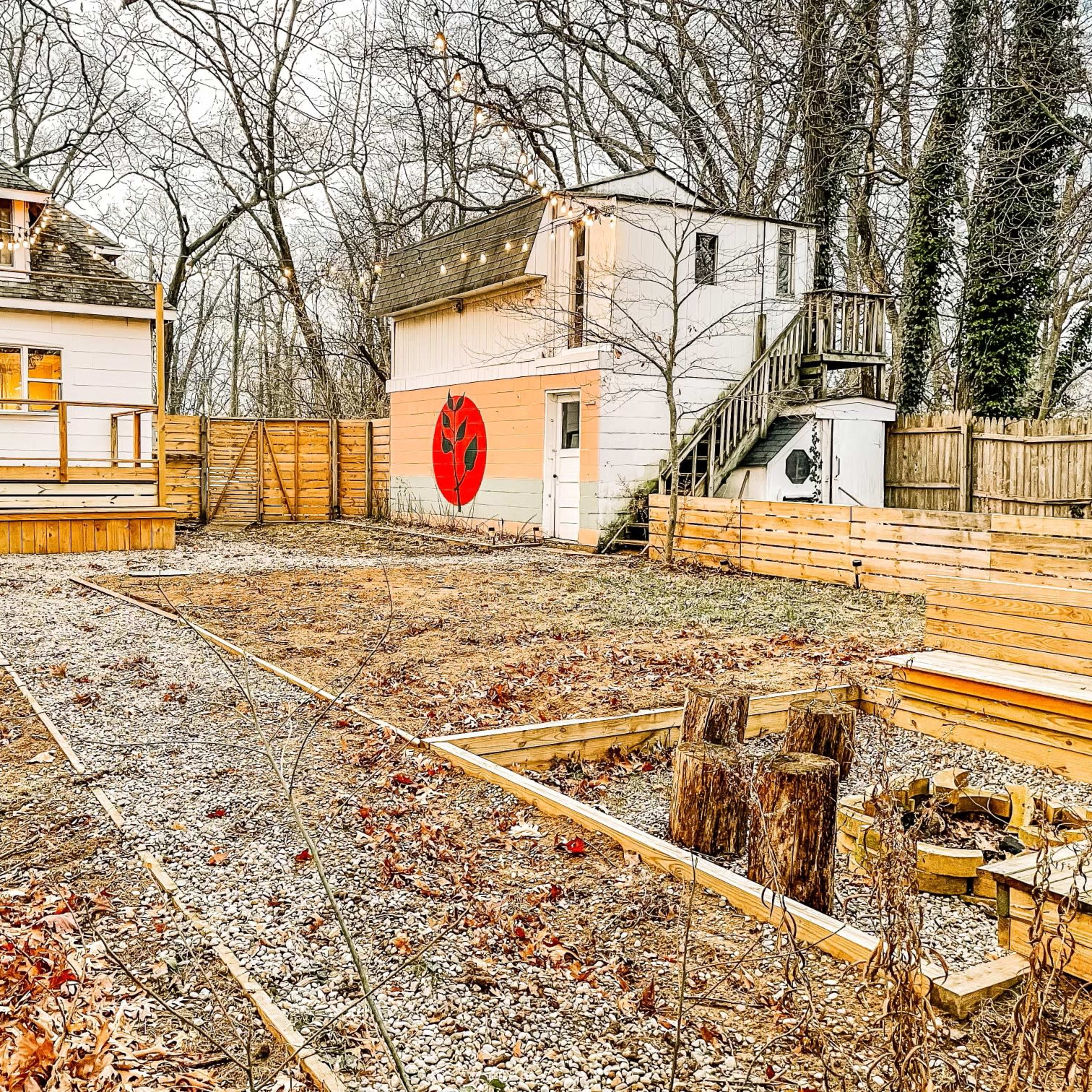 A vacant backyard with gravel and wooden borders, featuring two buildings, one with a red mural, and a set of logs arranged in a circle.