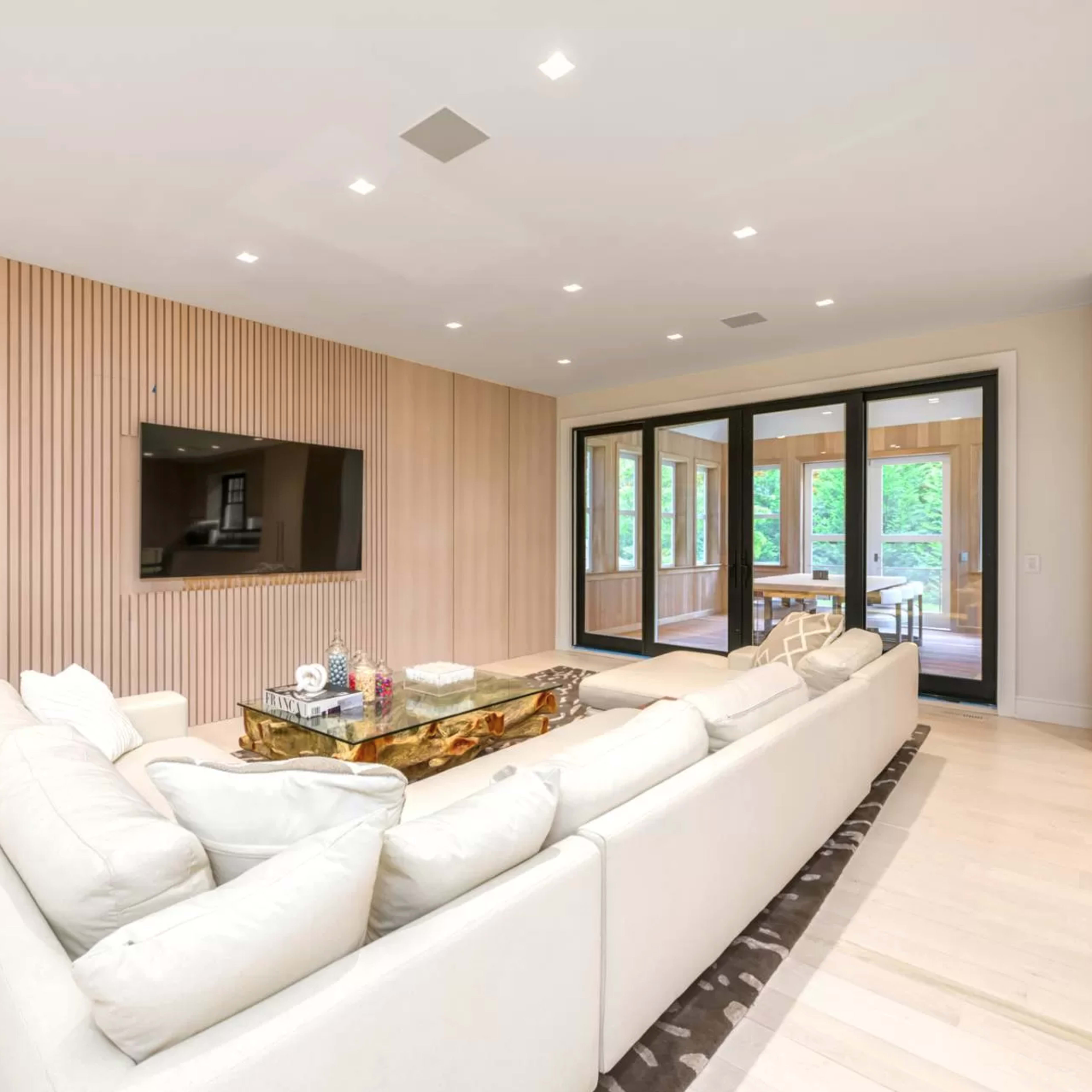 A modern living room features a white sectional sofa, a glass coffee table, and a large wall-mounted television against a wood-paneled backdrop.