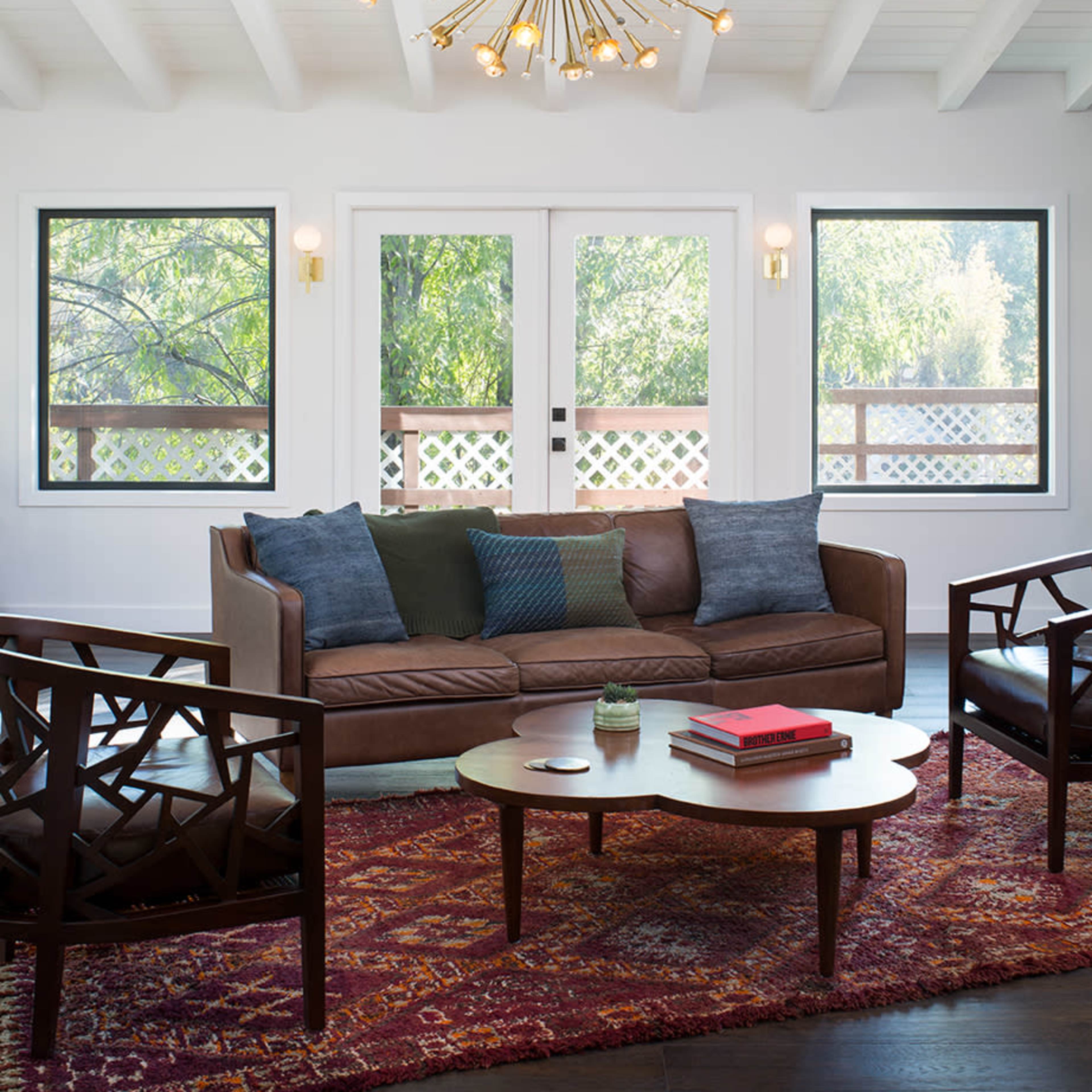 The living room features a brown sofa, two wooden chairs, a round coffee table, and large windows overlooking greenery, all illuminated by a starburst chandelier.
