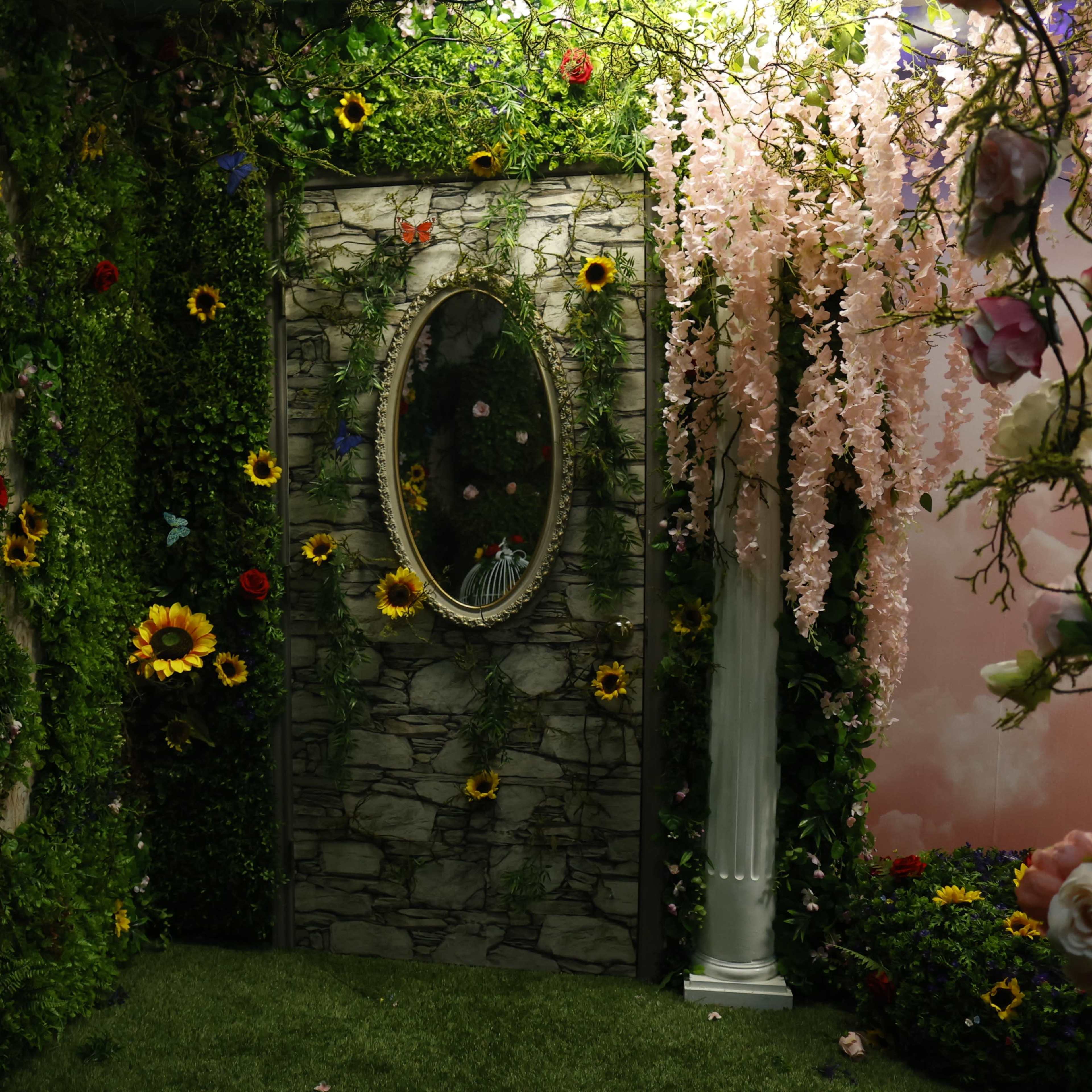 A decorative room with a stone wall adorned with various flowers and a mirror surrounded by foliage.