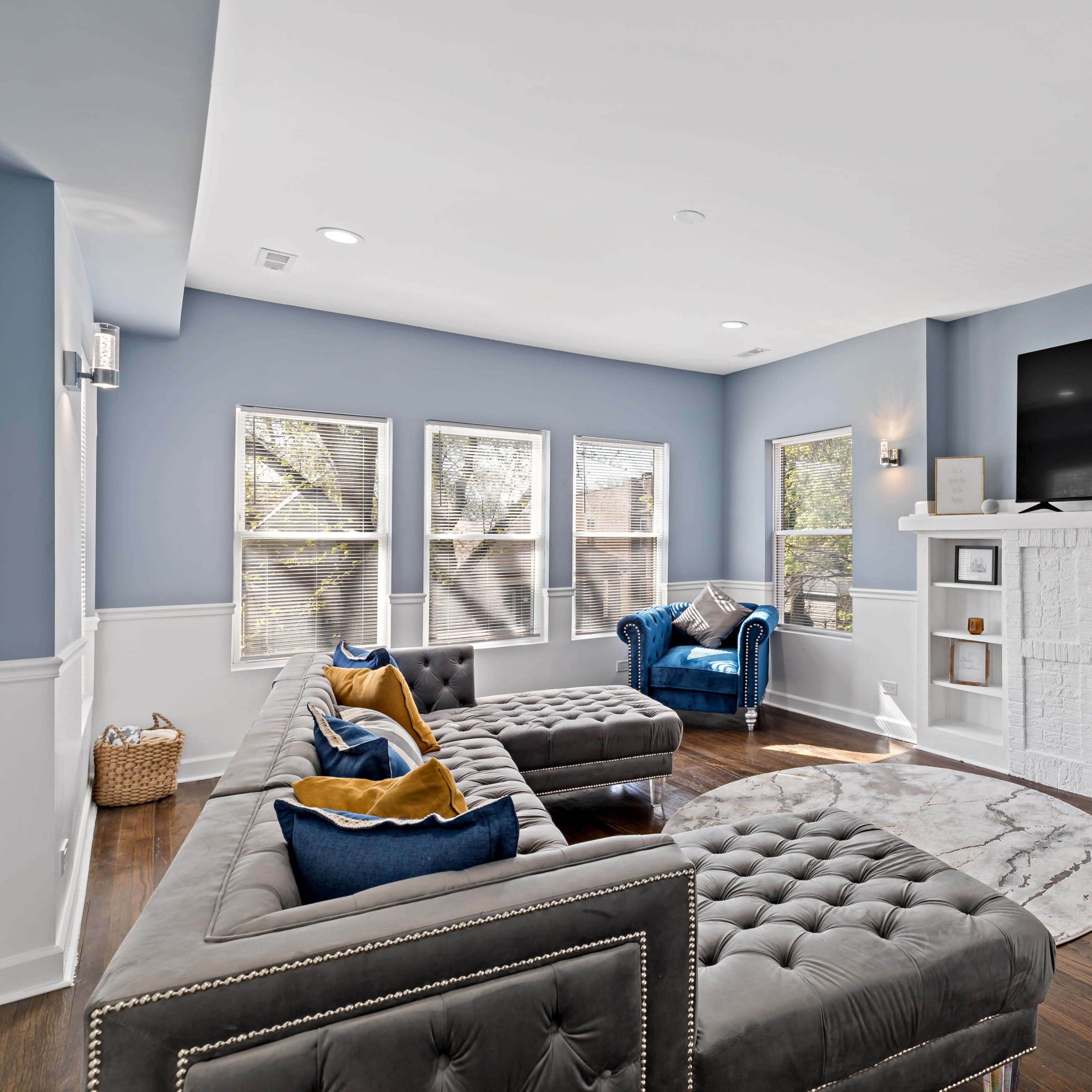 A spacious living room features a tufted sectional sofa, a round area rug, and a large television mounted above a white brick fireplace.