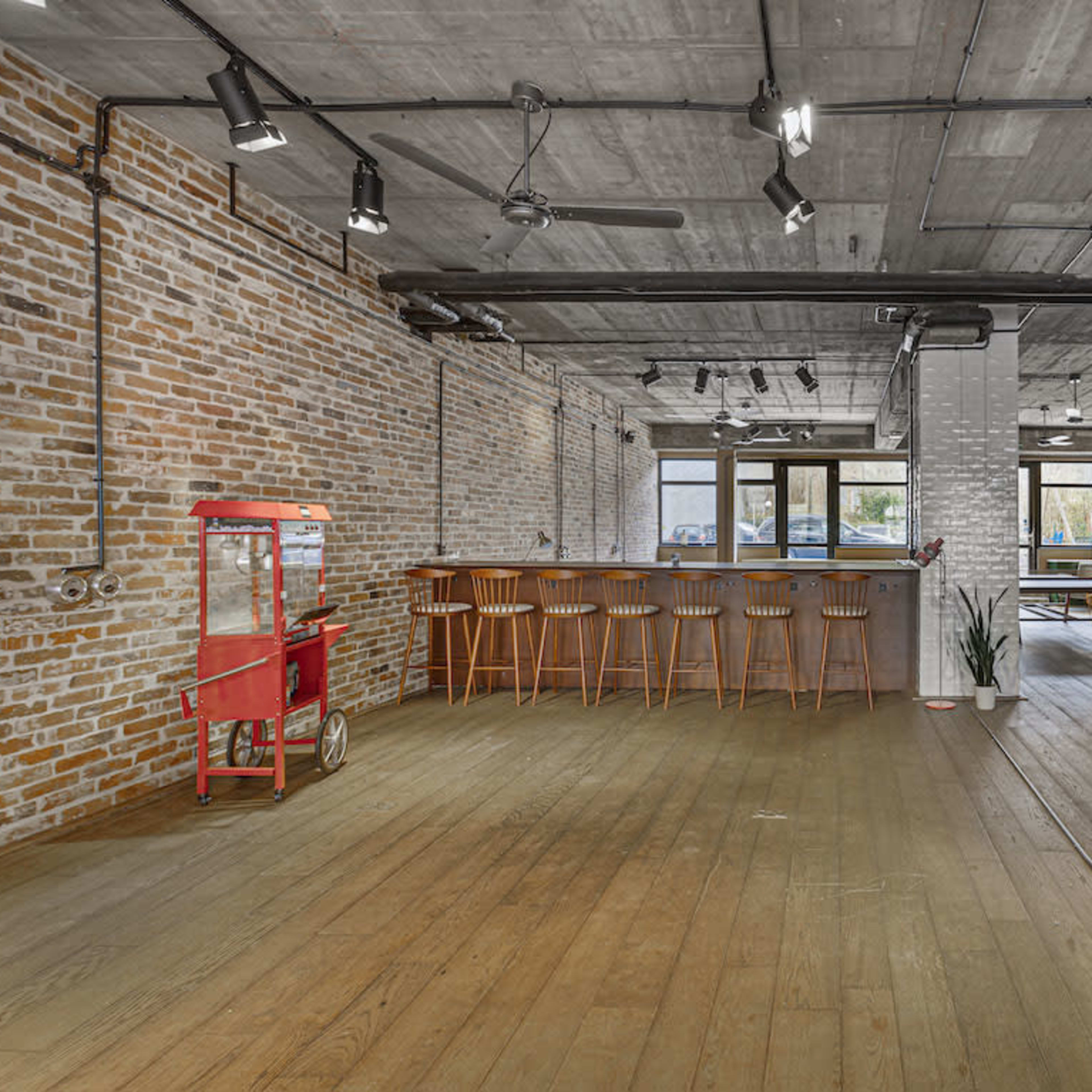 An open industrial-style interior space featuring exposed brick walls, wooden flooring, a bar area with stools, and a red cart in the corner.