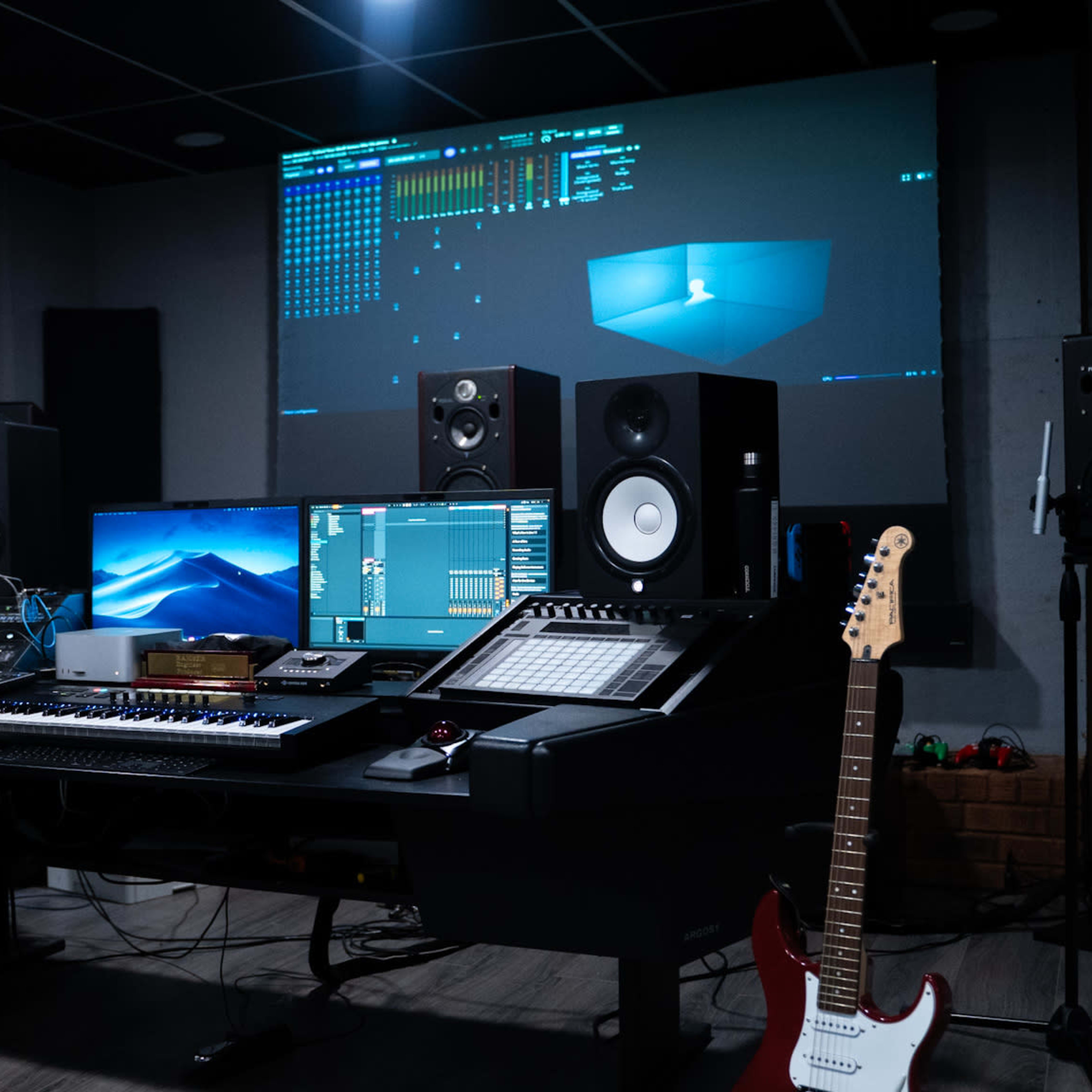 A music production studio features a large desk with multiple screens, speakers, a keyboard, and various equipment, along with a microphone and a guitar on the floor.