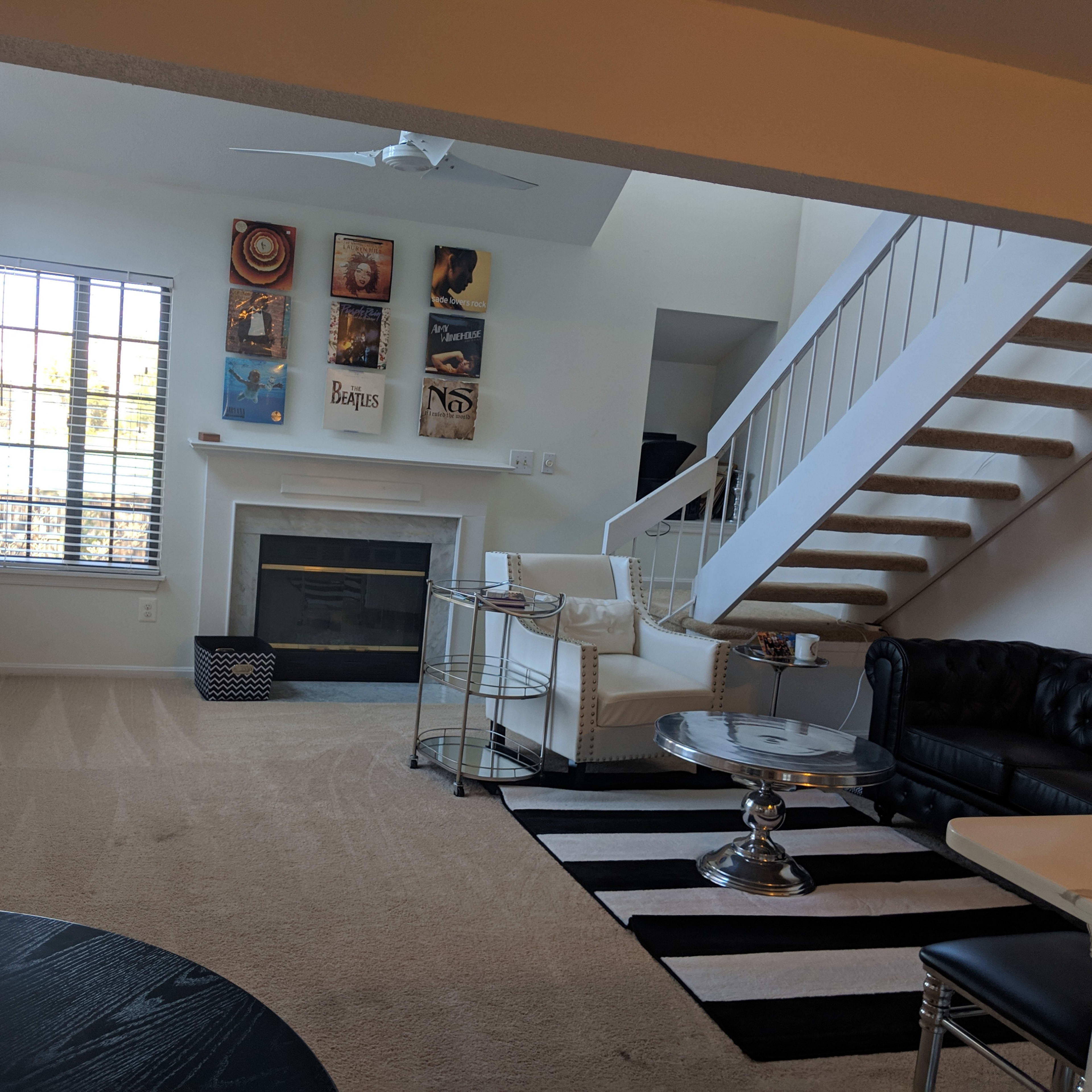 A living room with a fireplace, a black leather couch, a white sofa, a staircase, and a large window allowing sunlight to enter.