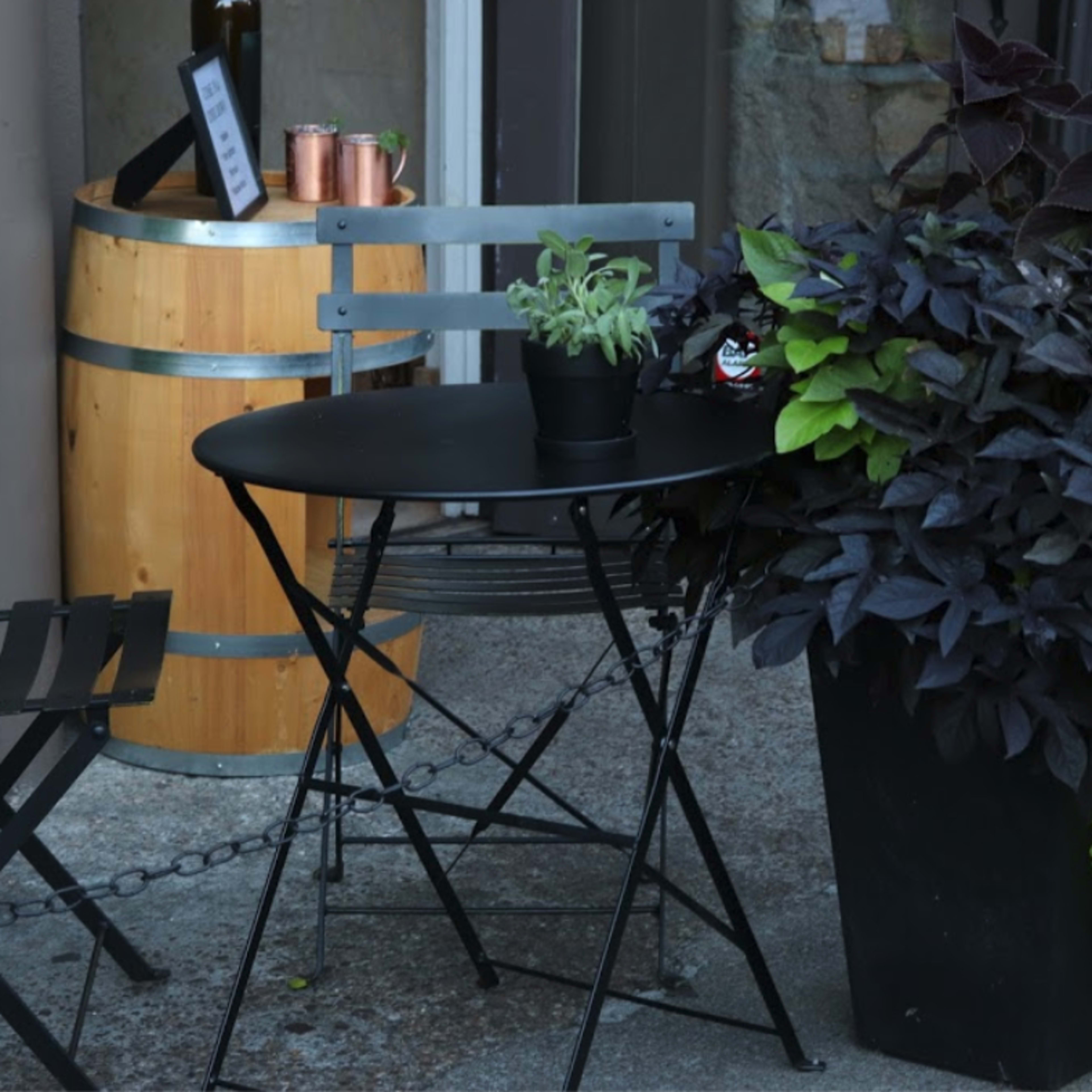 A small outdoor seating area features a black round table with a potted plant, two folding chairs, and a wooden barrel nearby.