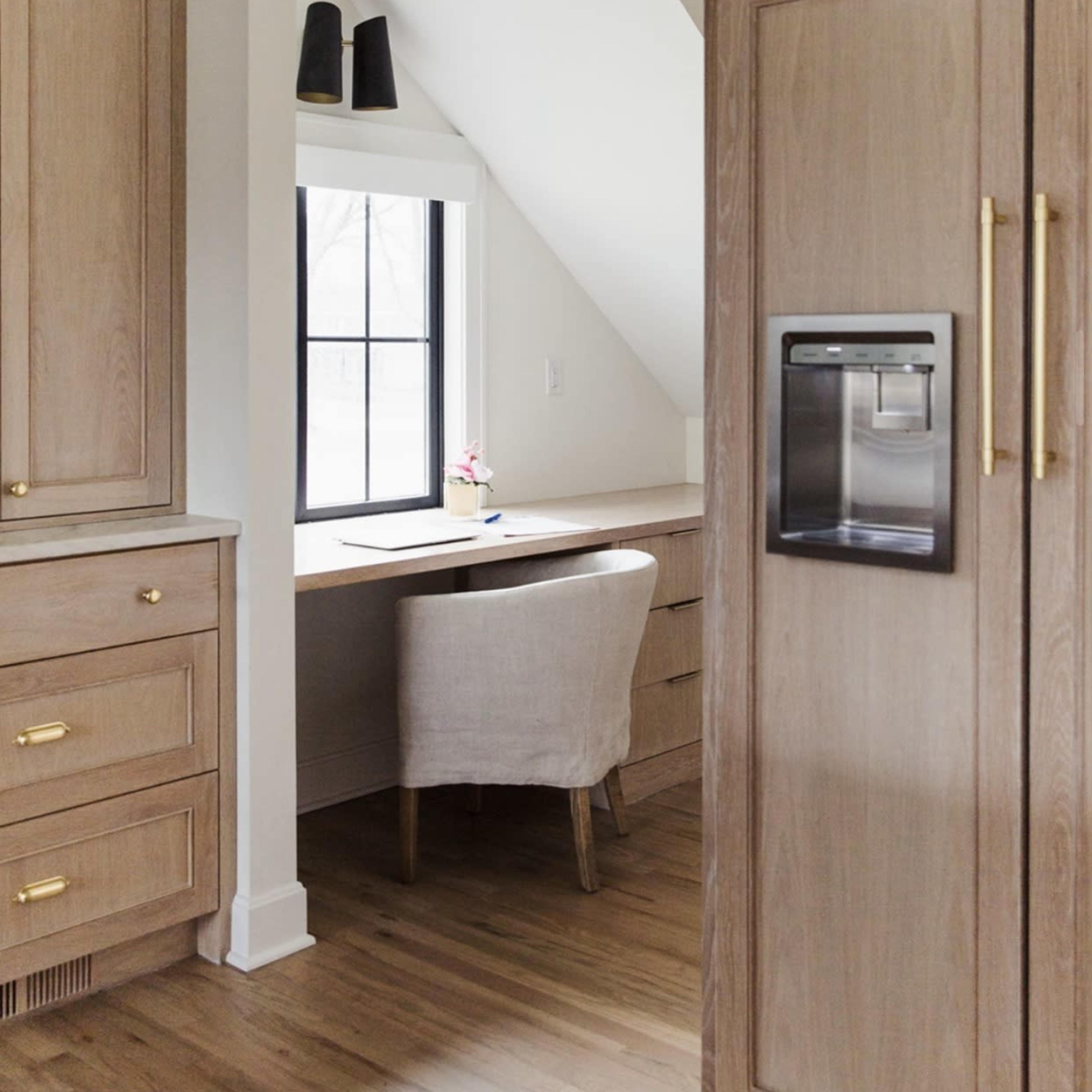 A small home office nook with a desk and a chair, located near a window, next to a modern kitchen featuring wooden cabinets and a refrigerator.