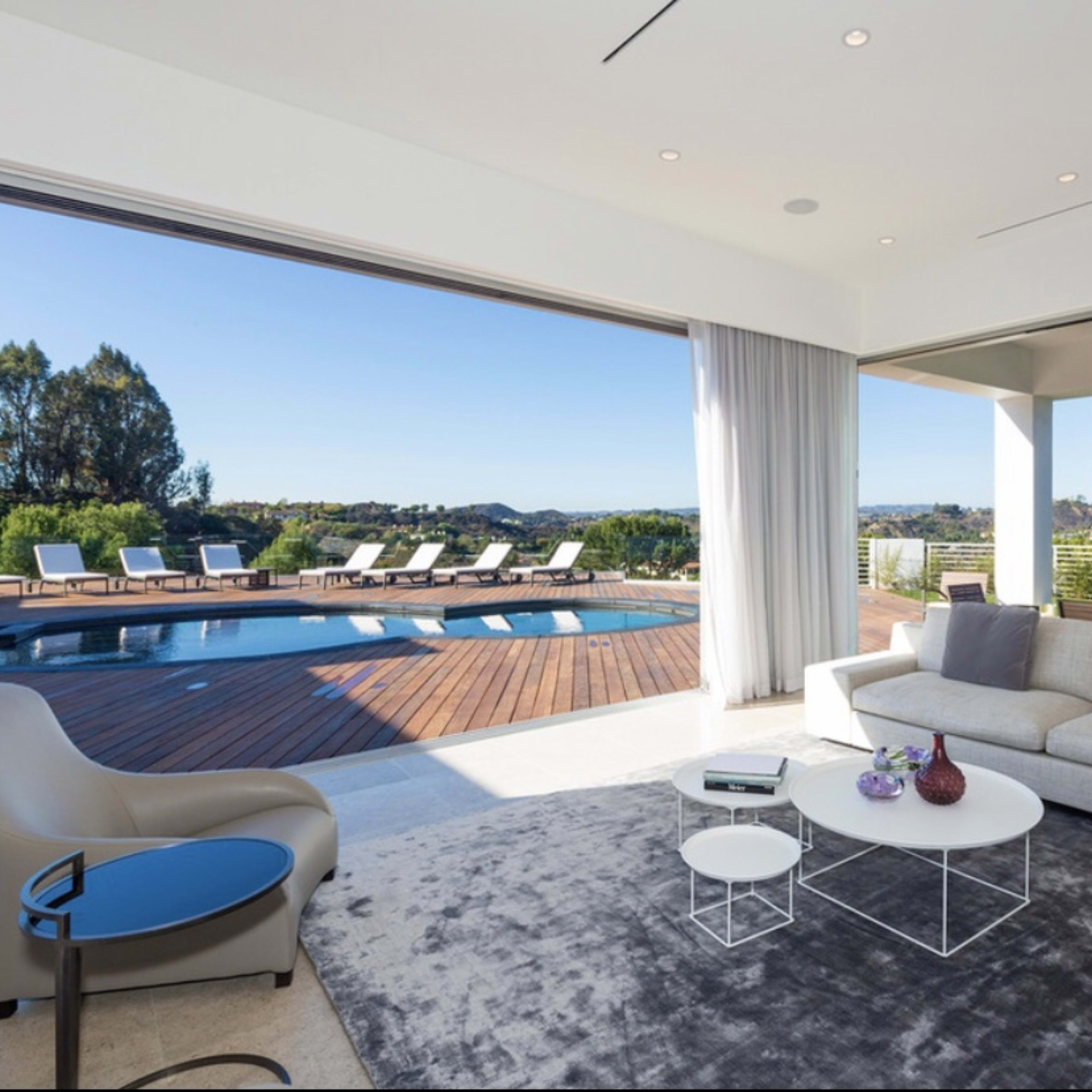 A modern living room features a large window that opens to a deck with a swimming pool and lounge chairs.
