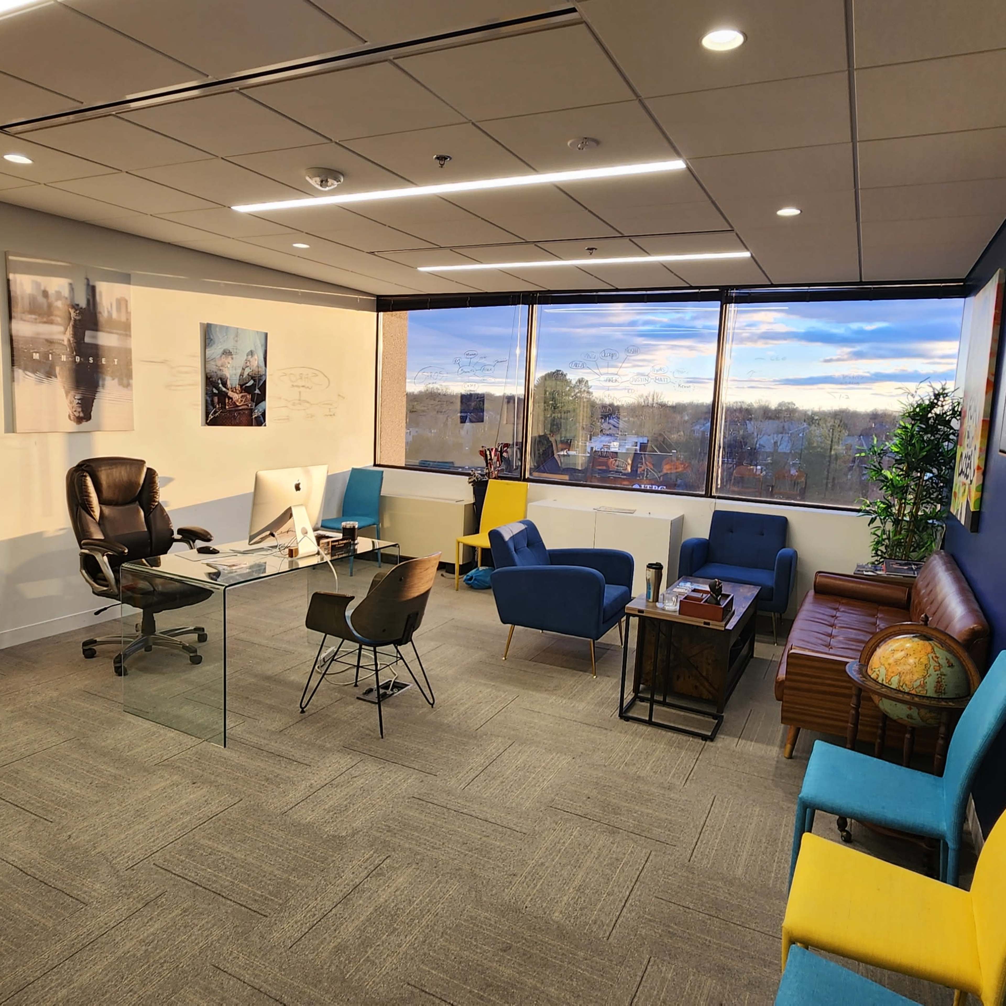 A modern office space featuring a glass desk with a black chair, various seating options in multiple colors, and large windows displaying a partially cloudy sky.