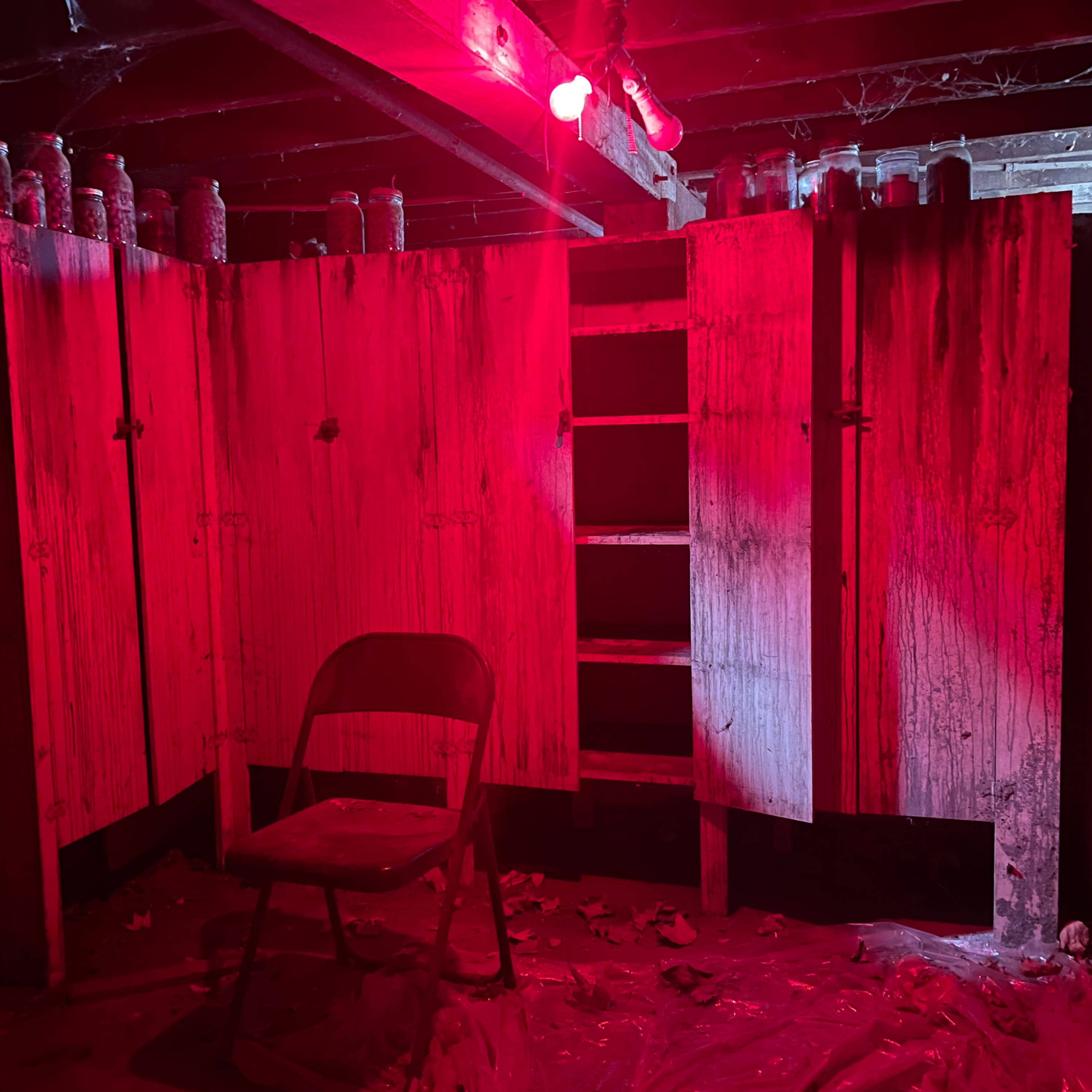 A dimly lit room with red lighting features a chair in front of weathered cabinets and jars on a shelf above.