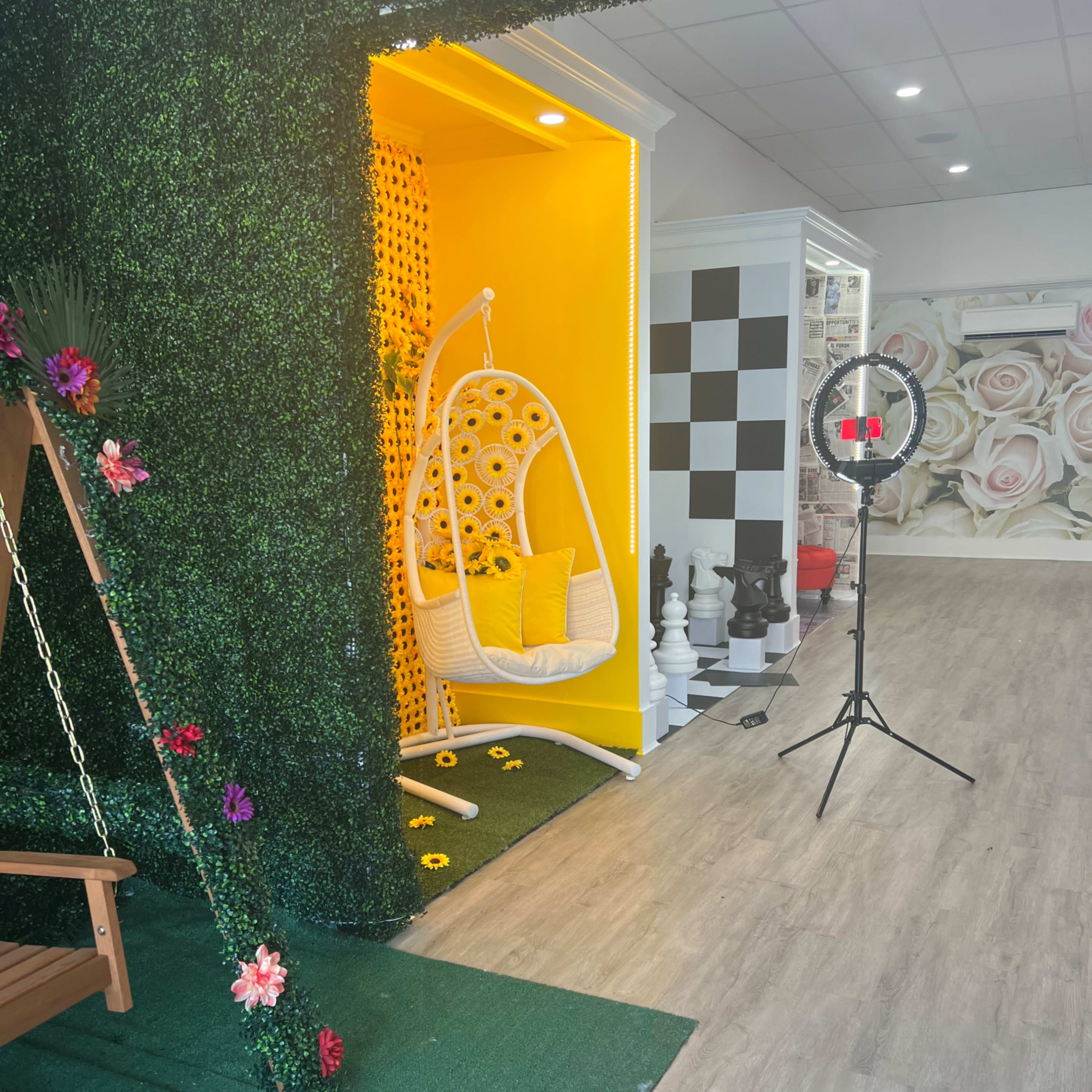 A brightly colored interior space featuring a yellow hanging chair surrounded by artificial greenery and a backdrop of pastel floral wallpaper.