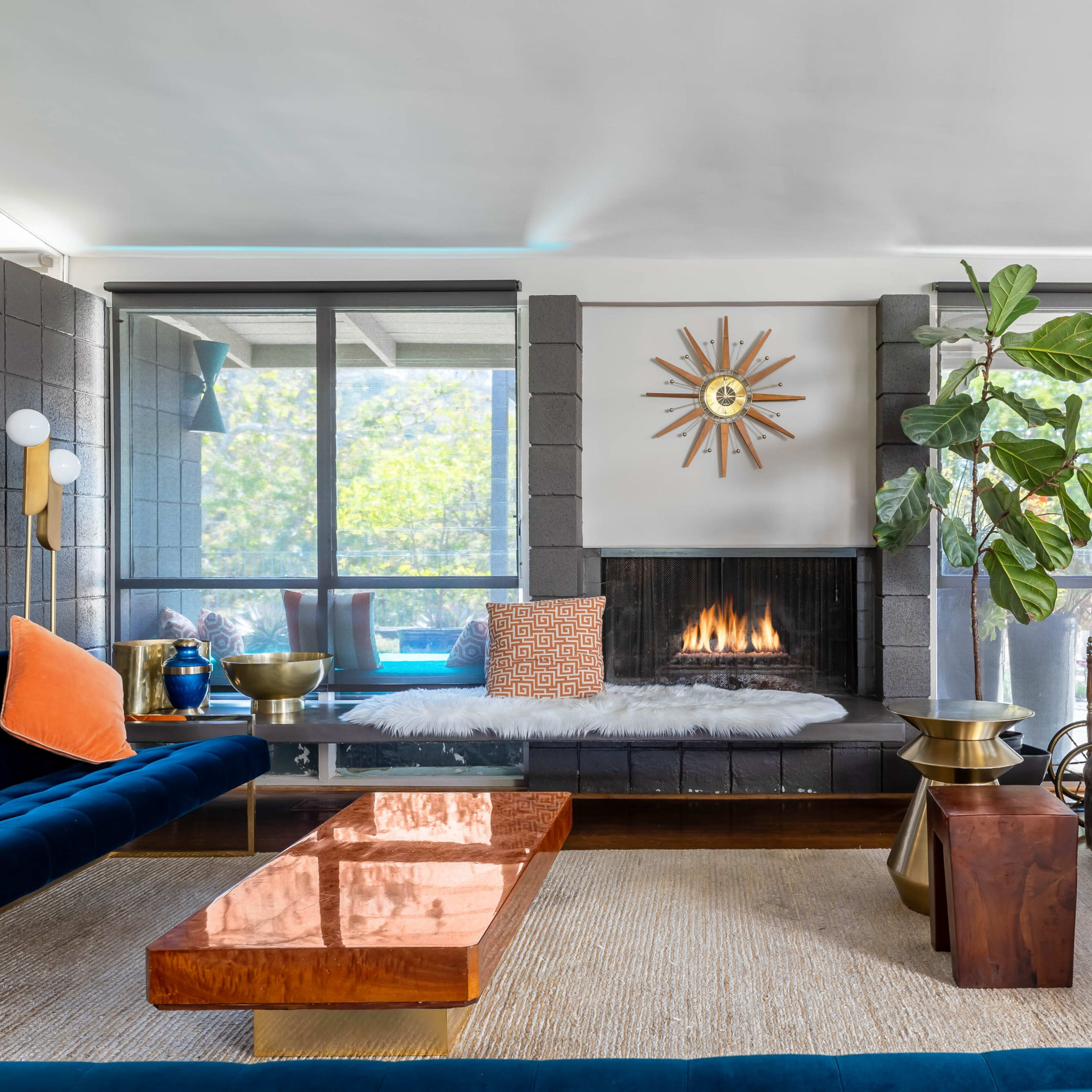 A modern living room features a fireplace, large windows, a blue velvet sofa, and a wooden coffee table.