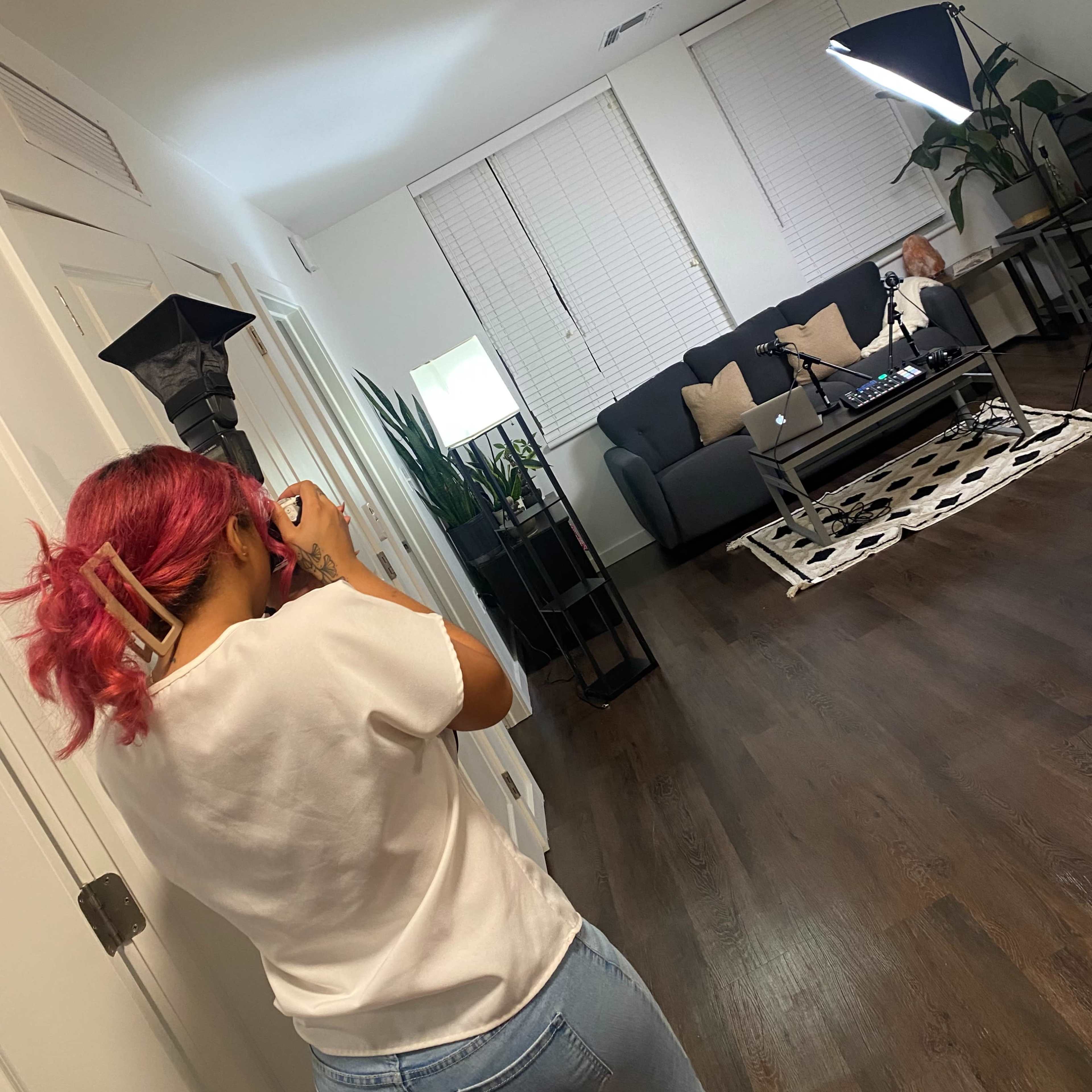 A person with pink hair takes a photo in a well-lit living room featuring a sofa, coffee table, and plants.
